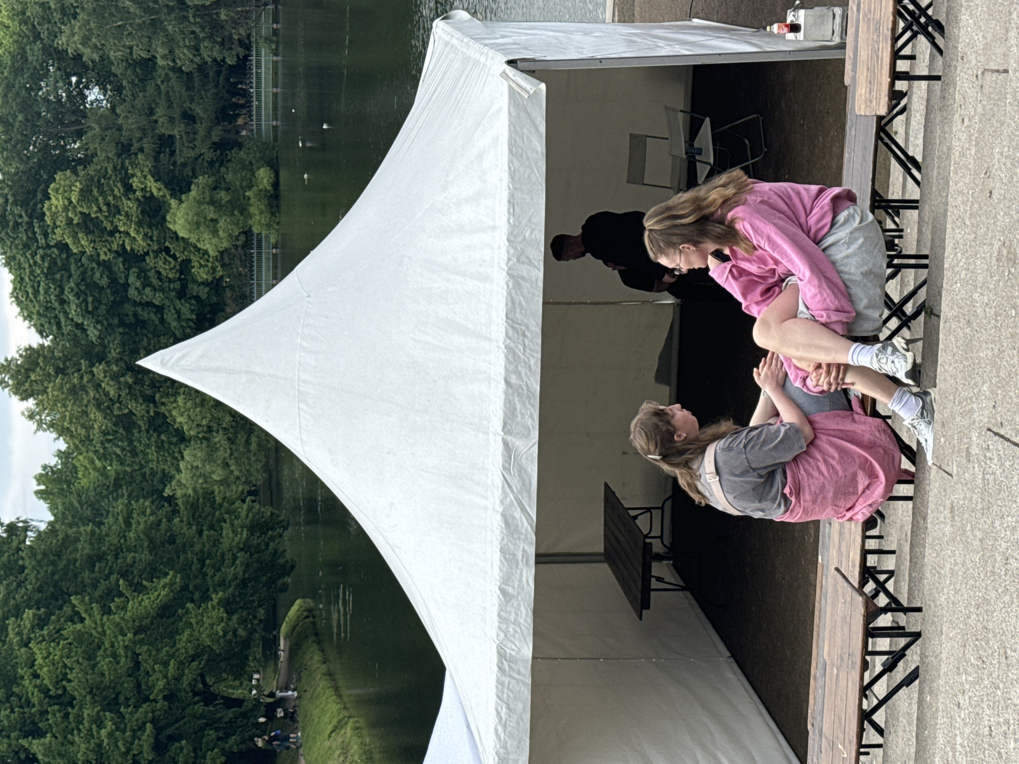 Two young women are sitting on a wooden bench in front of a large white tent. The woman on the left wears a grey t-shirt and a pink skirt. The woman on the right wears a pink hoodie and grey shorts. Both women are facing each other and appear to be in conversation. Inside the tent, a man is seated, and a table and chairs are visible. The tent is located outdoors, near a body of water and surrounded by lush green trees. The setting suggests a park or public outdoor space, possibly during a festival or event. The overall scene is one of casual interaction in a pleasant, natural environment.