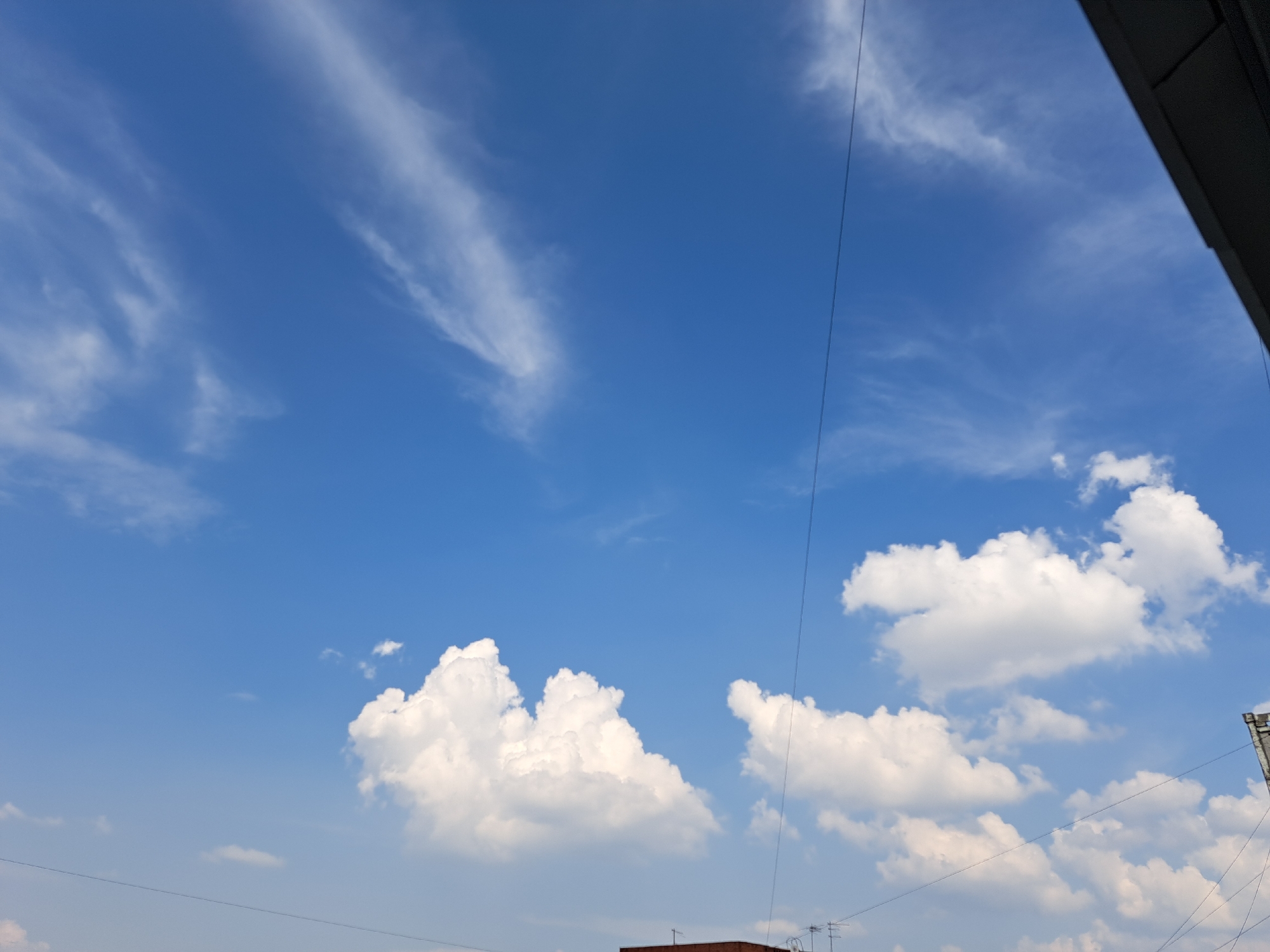 The image captures a daytime sky with scattered white clouds against a bright blue backdrop. A dark, angular structure, possibly part of a building or overhang, occupies the upper right corner. Thin, dark lines, likely electrical wires, traverse the sky. In the lower portion of the image, glimpses of rooftops and antenna-like structures suggest an urban environment. The scene depicts an outdoor setting in Irkutsk, Russia, without any visible people or specific actions taking place.