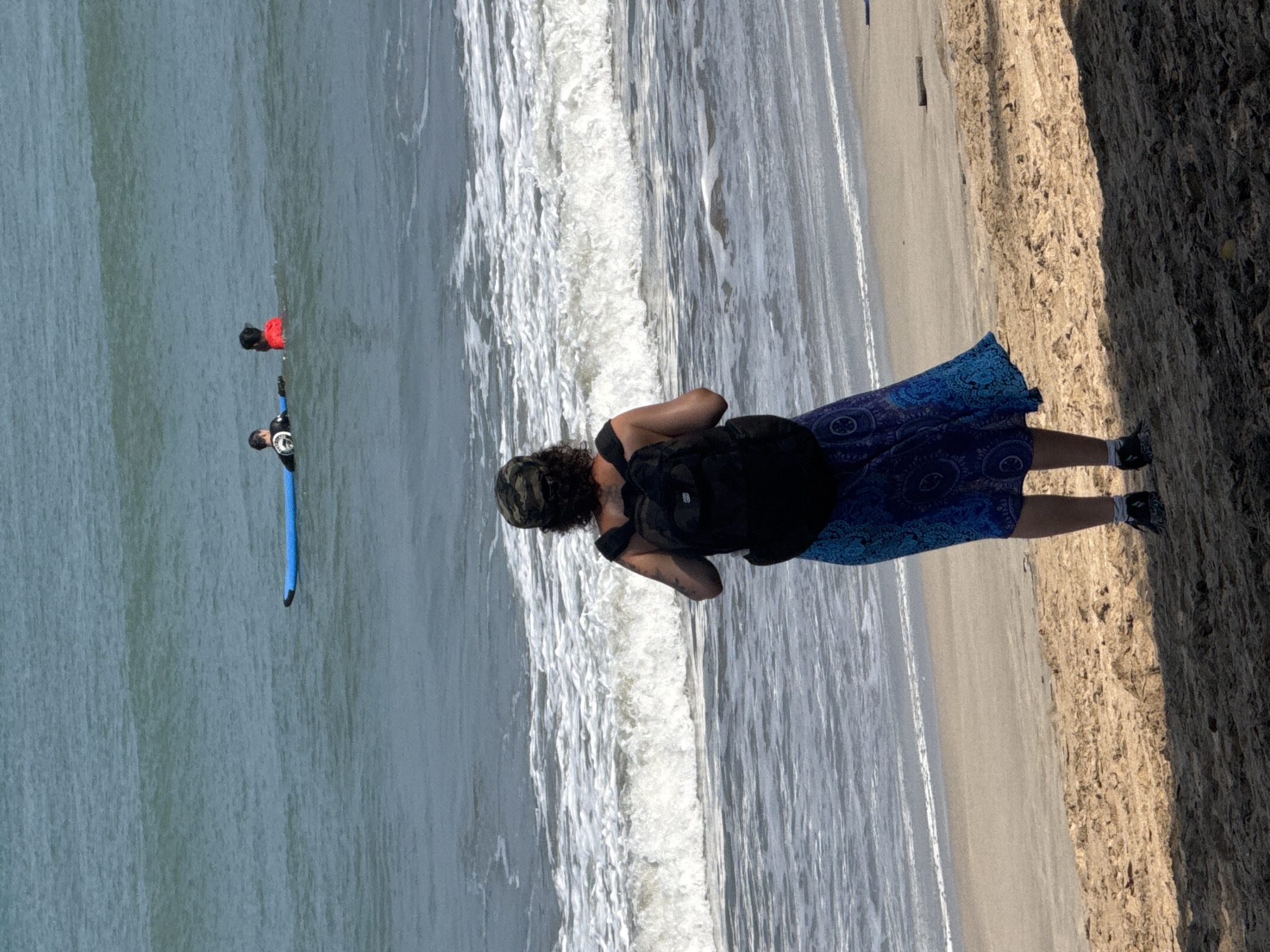A person with a black backpack and a patterned blue skirt stands on a sandy beach, facing the ocean. Two individuals are in the water. One is on a blue surfboard, and the other is in the water next to them. The ocean has gentle waves and a foamy white surf line. The beach is composed of sand with some debris. The sky is visible as a pale blue. The image appears to be taken in Kuta, Indonesia.