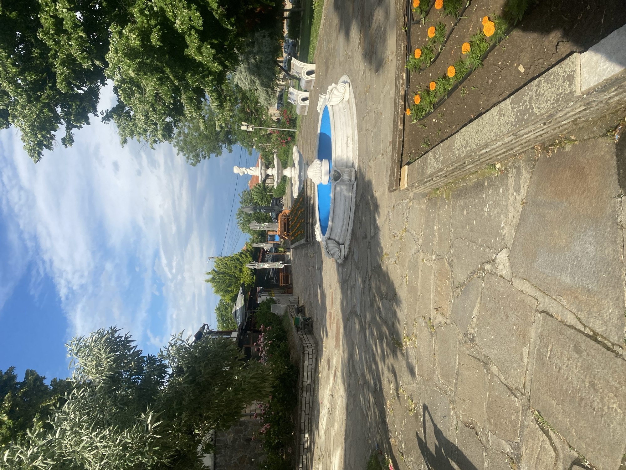 The image presents an outdoor scene in Ivaylovgrad, Bulgaria. A stone-paved path stretches into the distance, flanked by manicured garden beds on the right side, featuring rows of orange flowers. Along the path are several decorative white and blue fountains. Further down the path, white benches are visible. The surrounding area is filled with greenery, including trees and bushes. The sky is partially visible with blue and white skies.