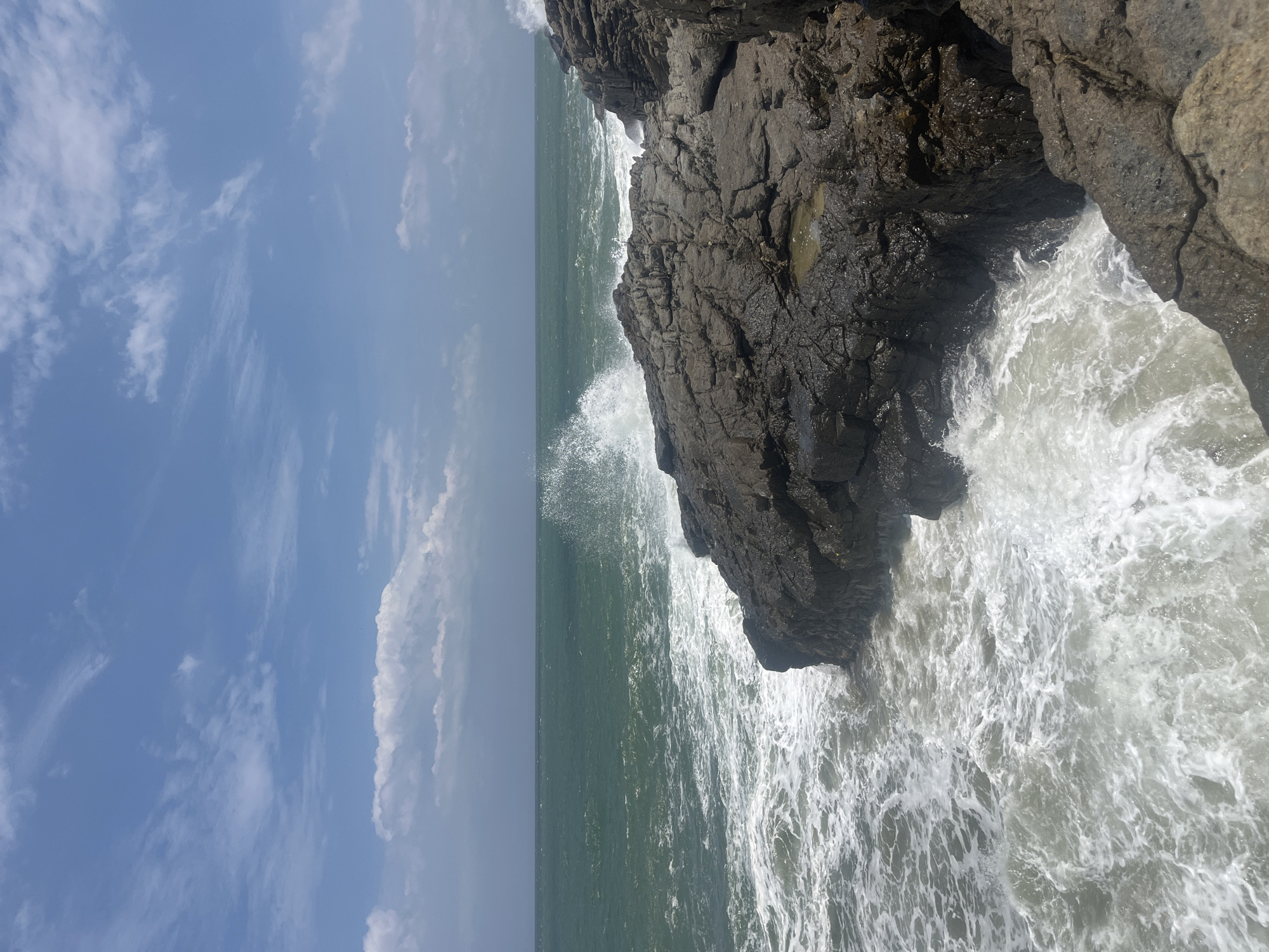 The image depicts a rocky coastline with waves crashing against the shore. The ocean is a deep green, and the sky is blue with scattered white clouds. The rocks are dark and appear wet, with some pools of water visible among them. A wave is breaking on the right side of the image, creating a splash of white foam. The scene is likely located in Mui Ne, Vietnam, a coastal town known for its beaches and natural beauty. There are no people or animals visible in the image. The overall impression is one of a rugged and wild coastline, with the power of the ocean on full display.