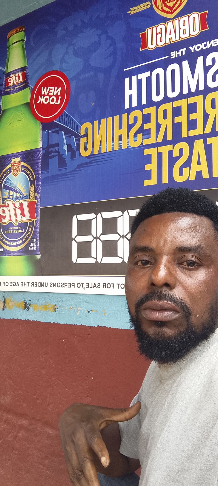 A man with a beard stands in front of a large advertisement for "Life" beer. The advertisement features a green bottle of Life beer, the brand logo, and text that reads "SMOOTH REFRESHING TASTE". There are also digital numbers displayed, possibly indicating a price or promotion. The background of the advertisement is blue with an image of a lion's head. The man is wearing a light grey t-shirt. His left hand is raised, with his index finger extended and pointing slightly upwards. The scene appears to be outdoors, with a brick wall in the foreground and a light blue painted surface above it. The advertisement is mounted on a wall. The location is identified as Gboko, Nigeria.
