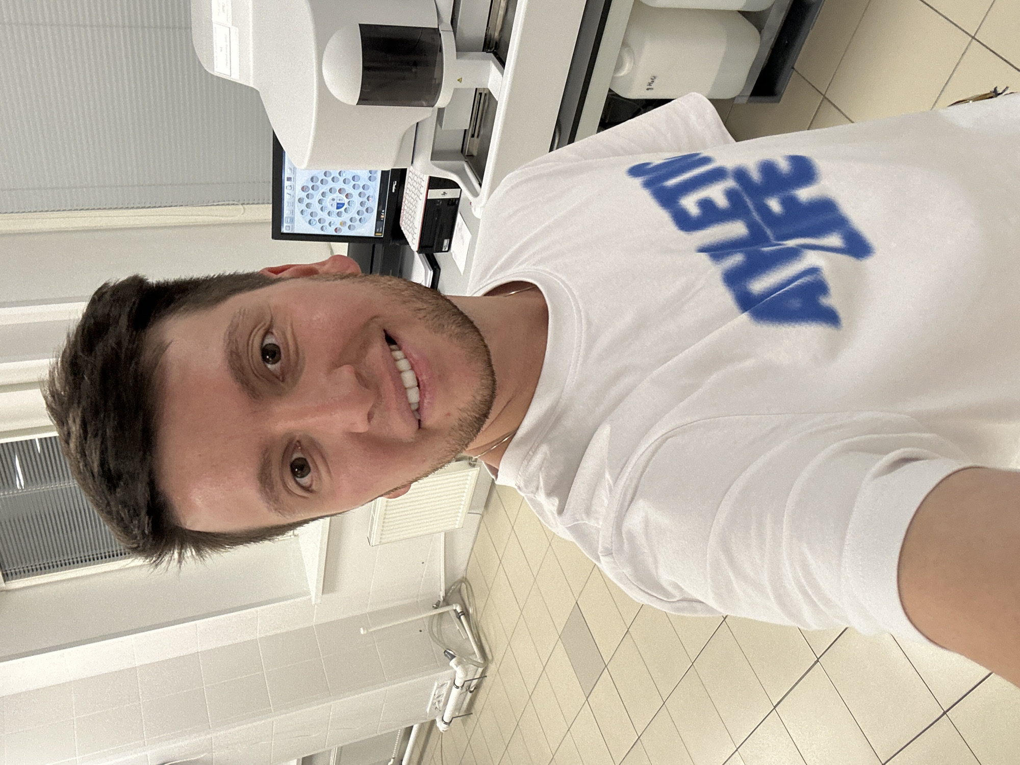 A young man with brown hair and a beard takes a selfie in what appears to be a laboratory. He is wearing a white t-shirt with blue text that reads "ATHLETIC LIFE." Behind him, a computer monitor displays what looks like a cell analysis program. To the right of the monitor is a piece of medical equipment, likely an analyzer. The room has tiled floors and walls, with a radiator visible on the left side. The overall impression is of a clinical or research environment.