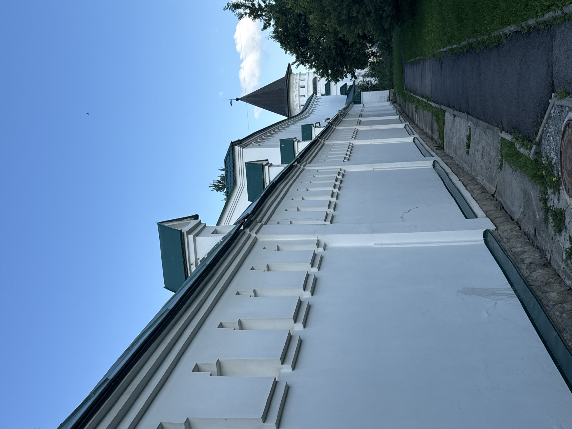 This image depicts an exterior view of a white building with a green roof, likely a historical or religious structure. The building features a distinct architectural style with stepped buttresses or decorative elements along its facade. A conical tower with a dark roof rises from the building, topped with a small weather vane. The building is situated on a slope, with a paved pathway running alongside it. Lush green grass and trees are visible in the background, suggesting a natural or park-like setting. The sky is a clear, bright blue with a few wispy clouds, indicating fair weather during the daytime. A small bird is visible in flight against the sky. The overall impression is one of a serene and possibly ancient location. The image appears to be taken from a low angle, looking up towards the building and the sky. The presence of a manhole cover with visible text on the right side of the image provides a location cue. The scene conveys a sense of tranquility and historical significance.