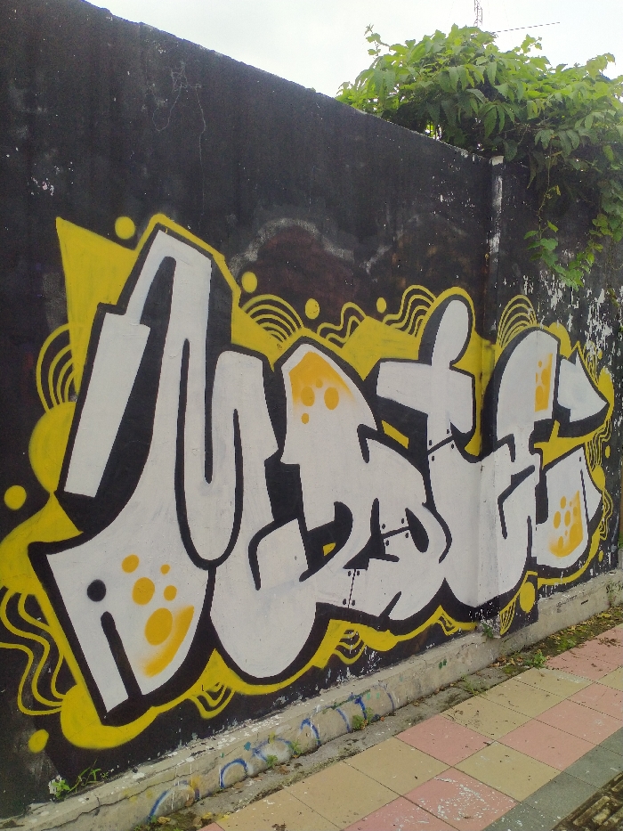 An outdoor shot captures a vibrant graffiti artwork spray-painted on a dark, weathered concrete wall. The wall, possibly a perimeter wall, is black and shows signs of age, with visible cracks and some discolored patches, particularly towards the top. Over the top edge and corner of the wall, lush green leafy vegetation is growing, suggesting an unkempt or naturalized environment. Above the wall, an overcast, light grey sky is visible, indicating daytime.
The prominent graffiti piece features large, bold, highly stylized letters outlined in black and filled with white paint. A bright yellow color is used both as an intricate background design, with wavy lines and circular accents, and as highlights within the white letters. Some white sections of the letters are further embellished with patterns of small yellow dots.
Upon closer inspection, the graffiti appears to spell the word "MUSTIKA." The letters are crafted in a complex wildstyle or bubble style, common in graffiti art, with interlocking and flowing elements. The first letter is a clear 'M', followed by a distinct 'U'. The subsequent letters, 'S', 'T', 'I', 'K', and 'A', are progressively more stylized but still discernible as part of the word.
At the base of the wall, a concrete curb or drainage channel runs parallel to the sidewalk. The sidewalk itself is paved with checkered tiles in alternating shades of pink and light beige or off-white. Some older, faded blue markings, possibly remnants of earlier graffiti or paint, are visible on the concrete curb. No people or active events are present in the static scene. The image provides a glimpse of urban or suburban street art in Karanganyar, Indonesia.
