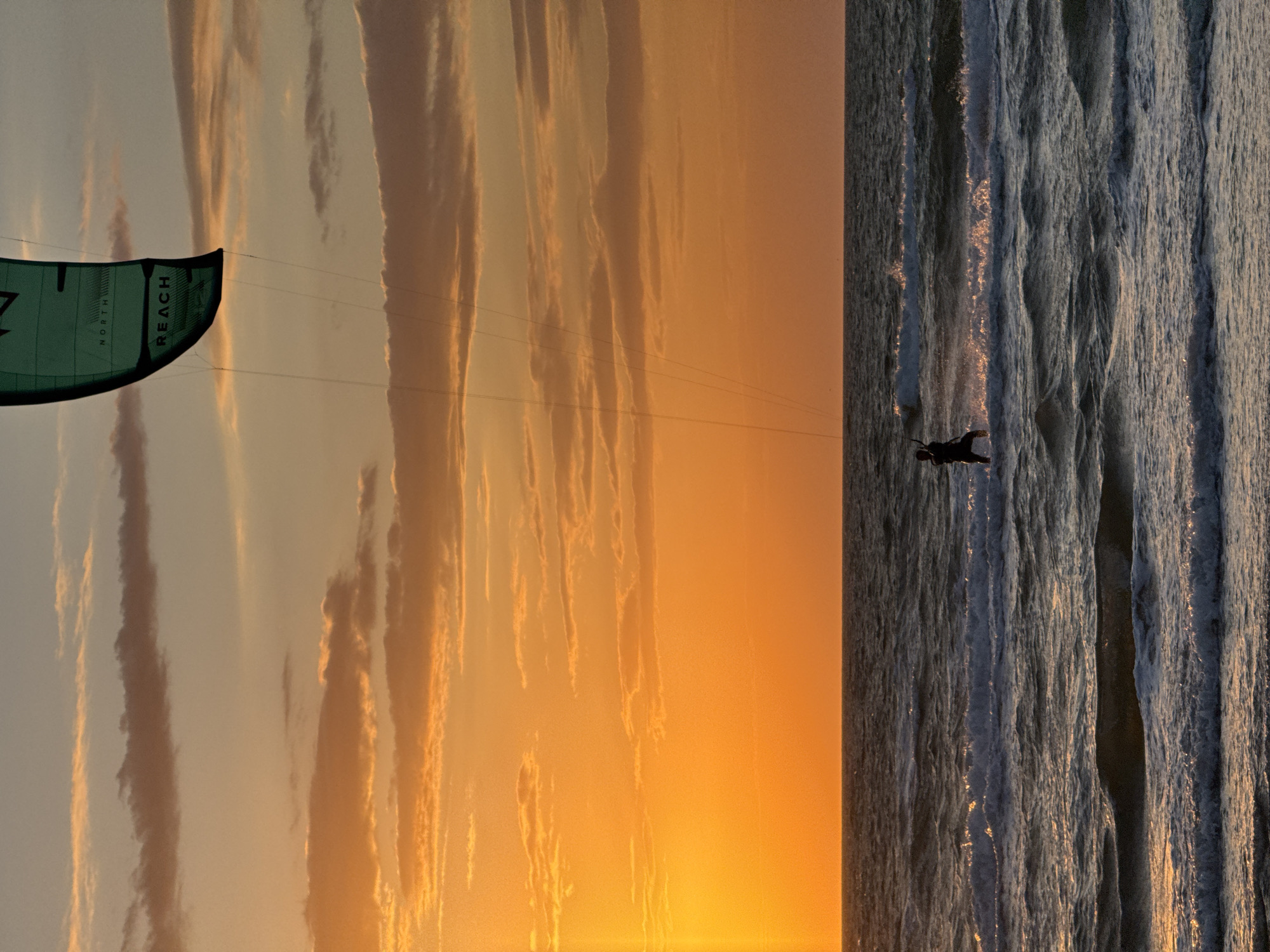 The image depicts a kitesurfer in the sea at sunset. The location is likely in the city of Shchelkino, Ukraine, given the prompt. The scene is divided, with a vibrant orange sky and striated clouds on the left, transitioning into the turbulent sea with visible waves on the right. The kitesurfer is a dark silhouette against the water. The sail, partially visible in the upper left, is labeled "REACH" and "NORTH."