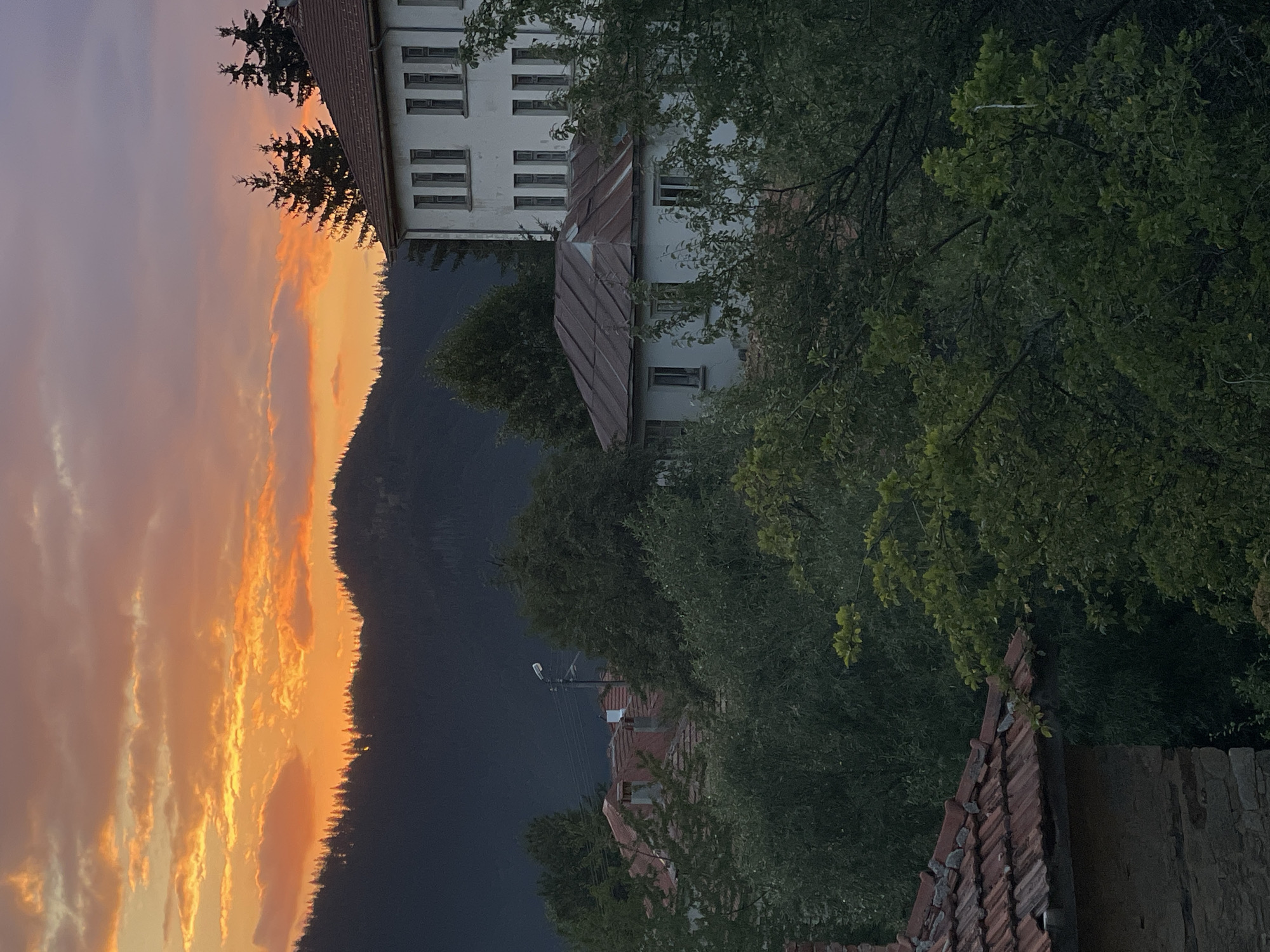 This is a photograph of a sunset over a village in Manastir, Bulgaria. The sky is filled with vibrant colors of orange, yellow, and pink, with clouds scattered throughout. Below the sky, a dark, tree-covered mountain range forms the horizon. In the foreground, several buildings with red tile roofs are visible, nestled amongst lush green trees. One larger, white building with several windows stands out to the right of the frame. The overall scene depicts a peaceful, rural setting at dusk.