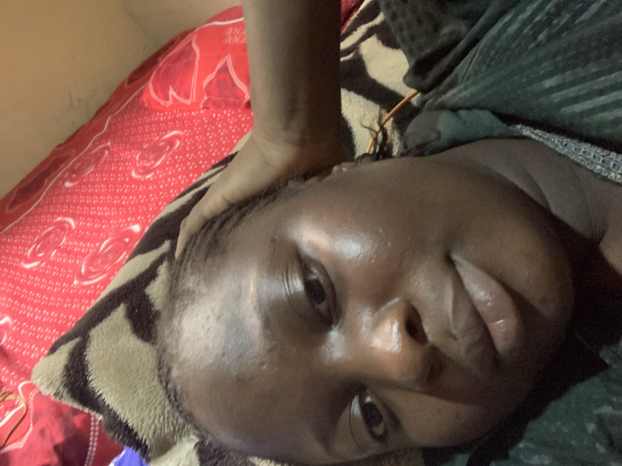 The image is a close-up, angled shot of a person lying on their back. The person's face is the primary focus, with their eyes closed and their mouth slightly open. A hand is visible resting on the person's head. The background consists of a red patterned fabric and a striped blanket. The lighting appears dim, suggesting an indoor setting. The overall impression is of a private, resting moment.