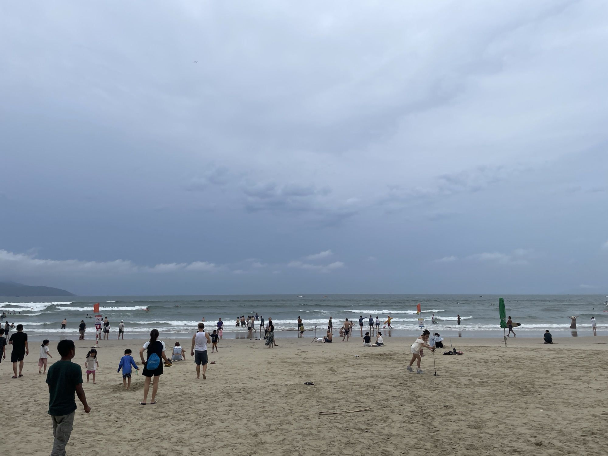 This image depicts a crowded beach scene at My Khe Beach in Da Nang, Vietnam, under an overcast sky. Numerous people are visible on the sandy shore and in the shallow waters. In the foreground, several individuals are walking or standing on the sand, including a group of children. Towards the water's edge, more people are engaged in various activities, some wading and others in the ocean where moderate waves are present. A lifeguard tower with a red flag is situated near the water, indicating a designated swimming area. The ocean stretches to the horizon, with a hazy mountain range visible in the distance. An airplane can be seen flying high in the sky. The overall atmosphere suggests a typical day at a popular beach with a mix of tourists and locals enjoying the seaside.