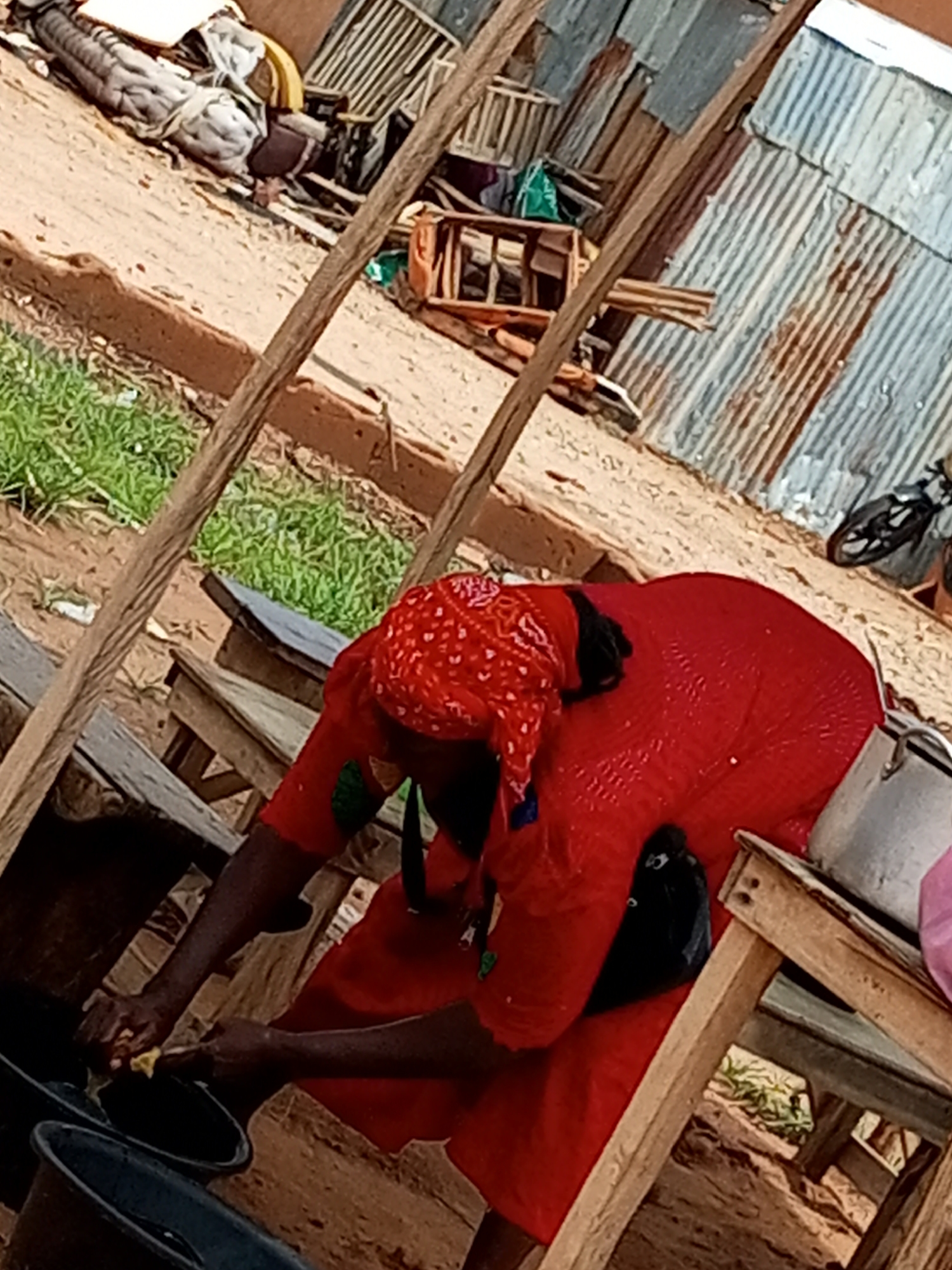 In Tempure, Nigeria, a woman in a red headscarf and red dress is washing dishes in black buckets on a wooden stall. Behind her, a corrugated metal structure serves as a wall. A motorcycle is parked in the background. The ground is sandy with some patchy green grass. Scattered debris, including wooden planks and what appears to be sacks, are visible.