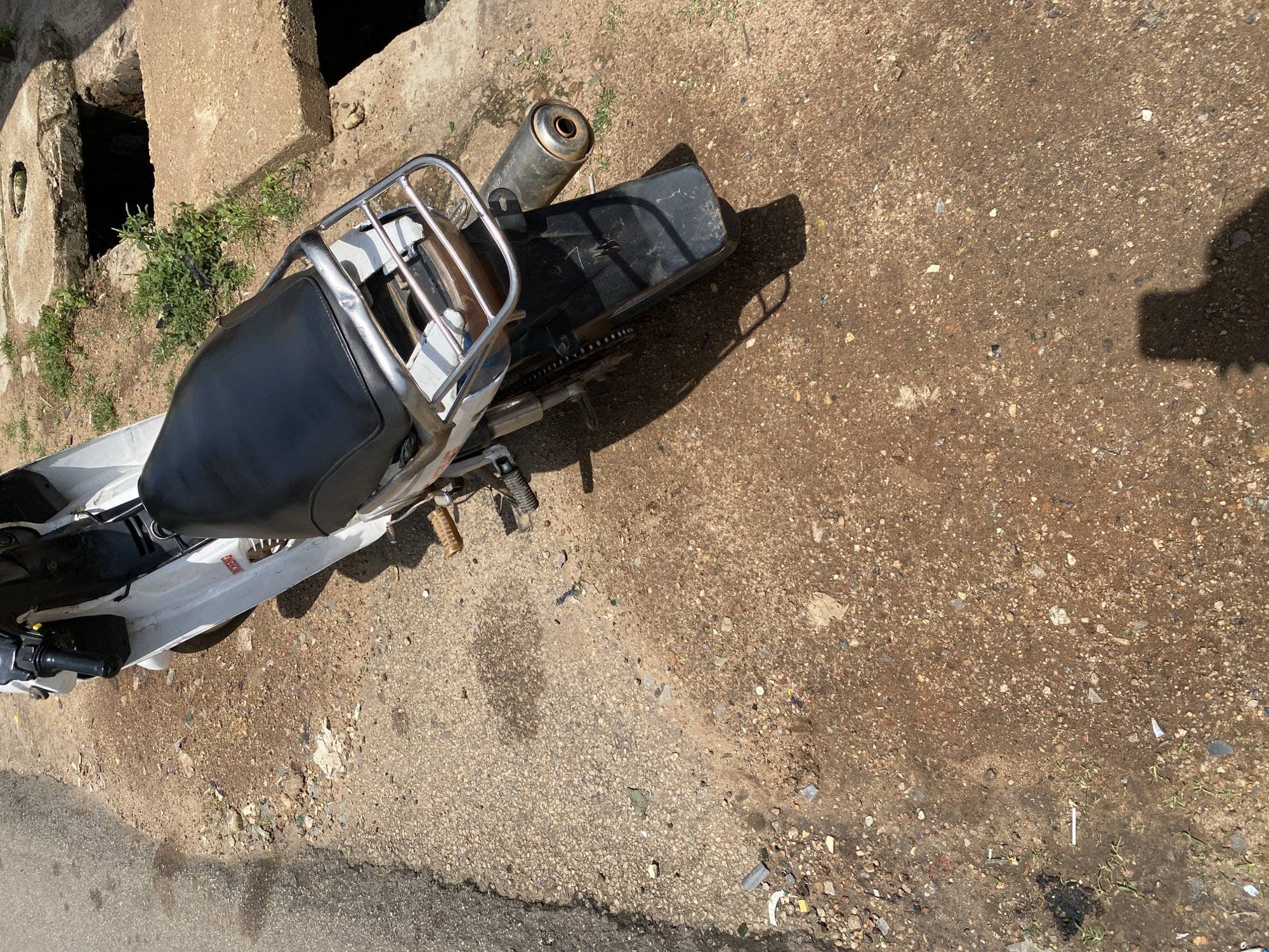 A white and black motorcycle is parked on a dirt and asphalt roadside in Jos, Nigeria. The motorcycle is partially visible, with its black seat, chrome rack, and exhaust pipe in focus. The ground is a mixture of reddish-brown dirt, small stones, and scattered debris, including plastic wrappers and cigarette butts. To the right of the motorcycle, there are concrete structures with open drainage channels. The scene is illuminated by sunlight, casting a shadow of a person on the ground in the lower right portion of the image.