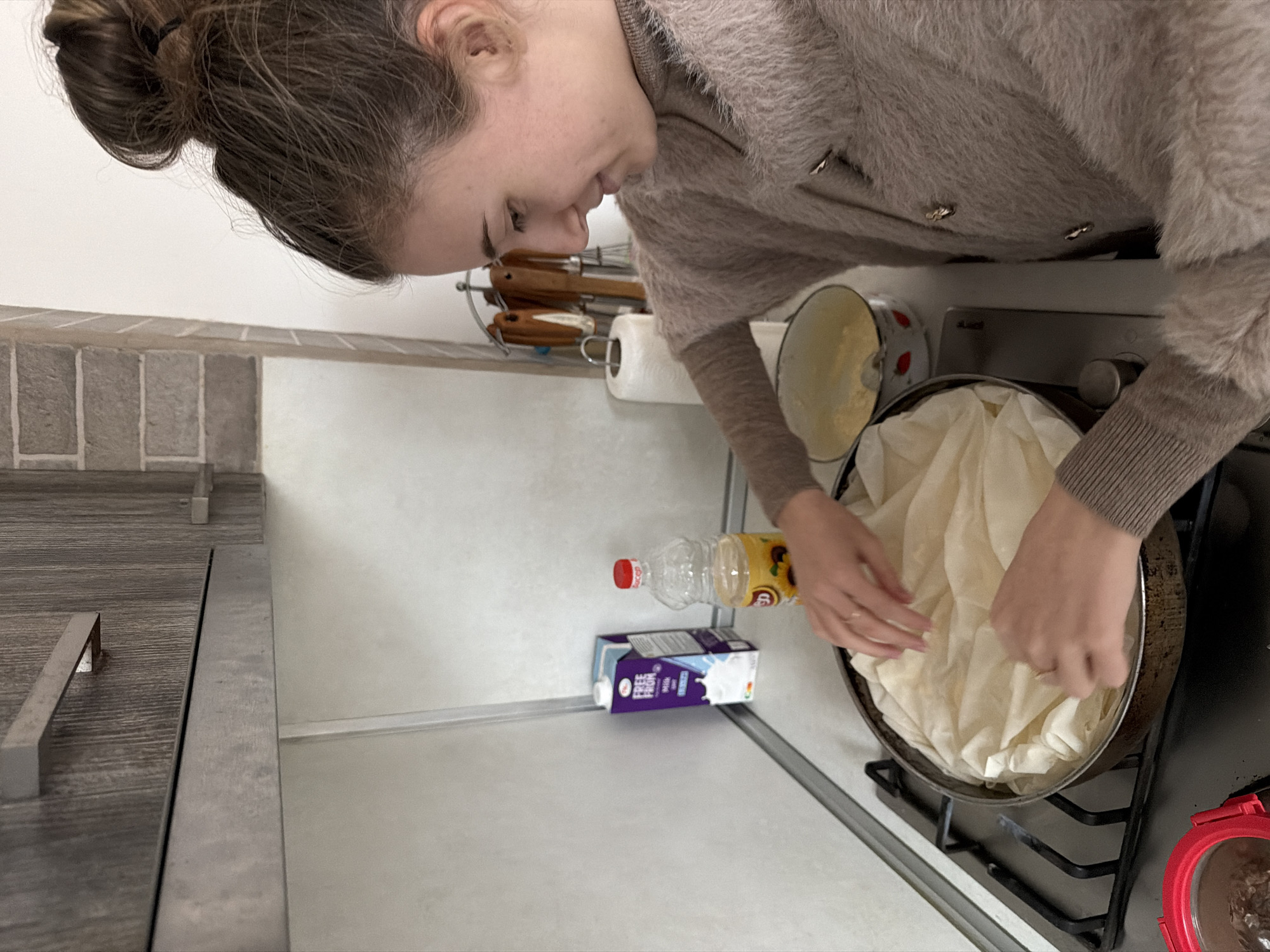 In an indoor kitchen setting, a person with brown hair pulled up into a bun and wearing a fuzzy, taupe cardigan is preparing food in a frying pan on a gas stovetop. The person is adding sheets of white phyllo dough to the pan. A bottle of oil and a carton of milk are on the countertop nearby. Kitchen utensils are hanging on a rack in the background. The specific location is not discernible from the image.