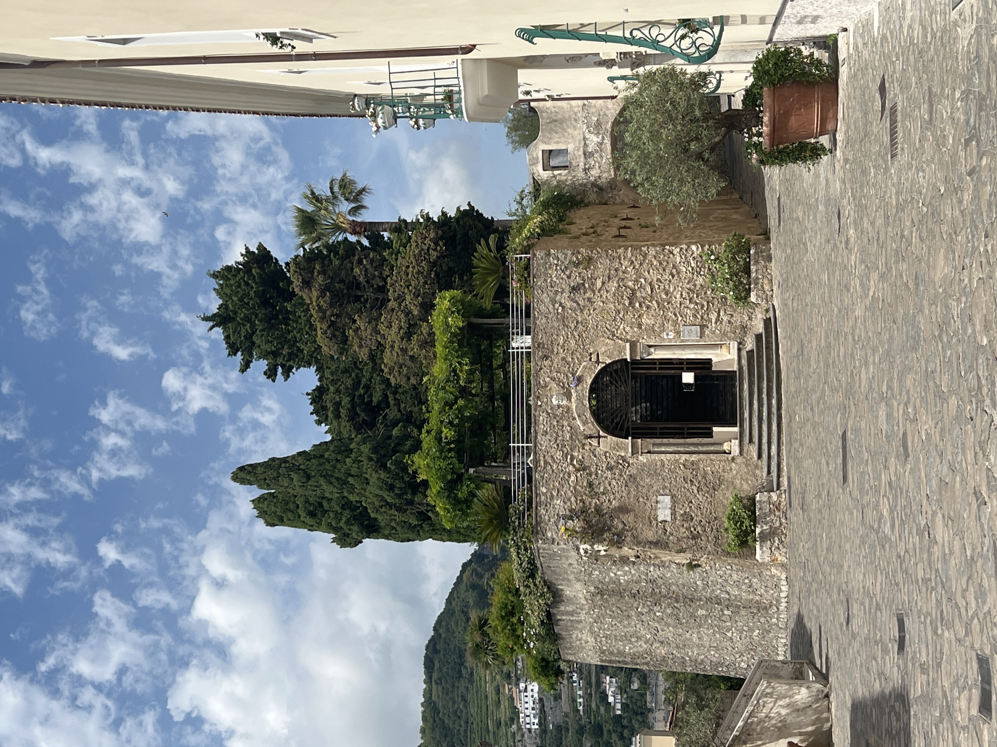The image shows an outdoor scene in Ravello, Italy. In the foreground, a cobblestone plaza leads to a stone structure with an arched entrance. The entrance has a dark metal gate and is flanked by steps. Above the entrance, a balcony with a white railing is visible, adorned with greenery. A mature olive tree in a large terracotta pot is positioned to the right of the stone structure, with its branches cascading over the side. Behind the stone structure, lush vegetation, including cypress trees and a palm tree, grows on a terraced hillside. In the background, a light-colored building with a balcony featuring decorative railings and potted plants stands to the right. The sky is blue with scattered white clouds. The overall impression is of a tranquil, scenic courtyard in a historic Italian town.