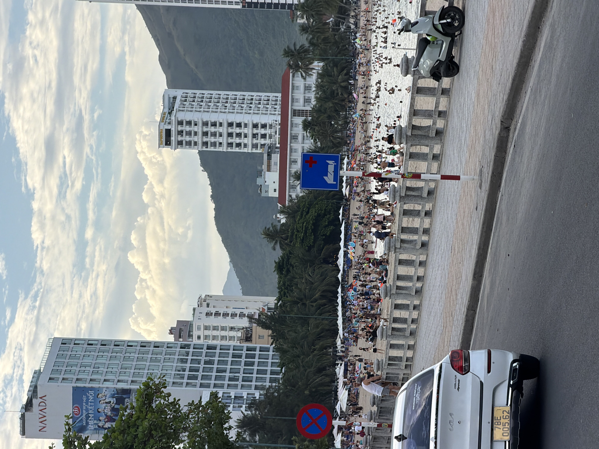 The image depicts a bustling beach scene in Nha Trang, Vietnam, likely along Tran Phu Beach, a popular tourist destination. In the foreground, a white car is parked on the right side of a road. To the left of the car, a concrete sidewalk lines the road, leading to a grey stone wall that separates the promenade from the beach.
Beyond the wall, a crowded beach stretches out, filled with people relaxing on lounge chairs and umbrellas, swimming in the ocean, and strolling along the sand. Several tall, modern buildings, characteristic of Nha Trang's coastal skyline, are visible in the background. One prominent building on the left features a large advertisement banner with Vietnamese text and images of people. Another tall white building, possibly a hotel, is situated further down the coast. Mountains form a backdrop to the city.
Traffic signs are present, including a blue and red circular sign prohibiting parking on the left and a blue rectangular sign with a white cross indicating a first-aid station. A light green scooter is parked on the sidewalk. The sky is partly cloudy with the sun shining through, casting a soft light on the scene. The overall atmosphere is one of a lively holiday destination.