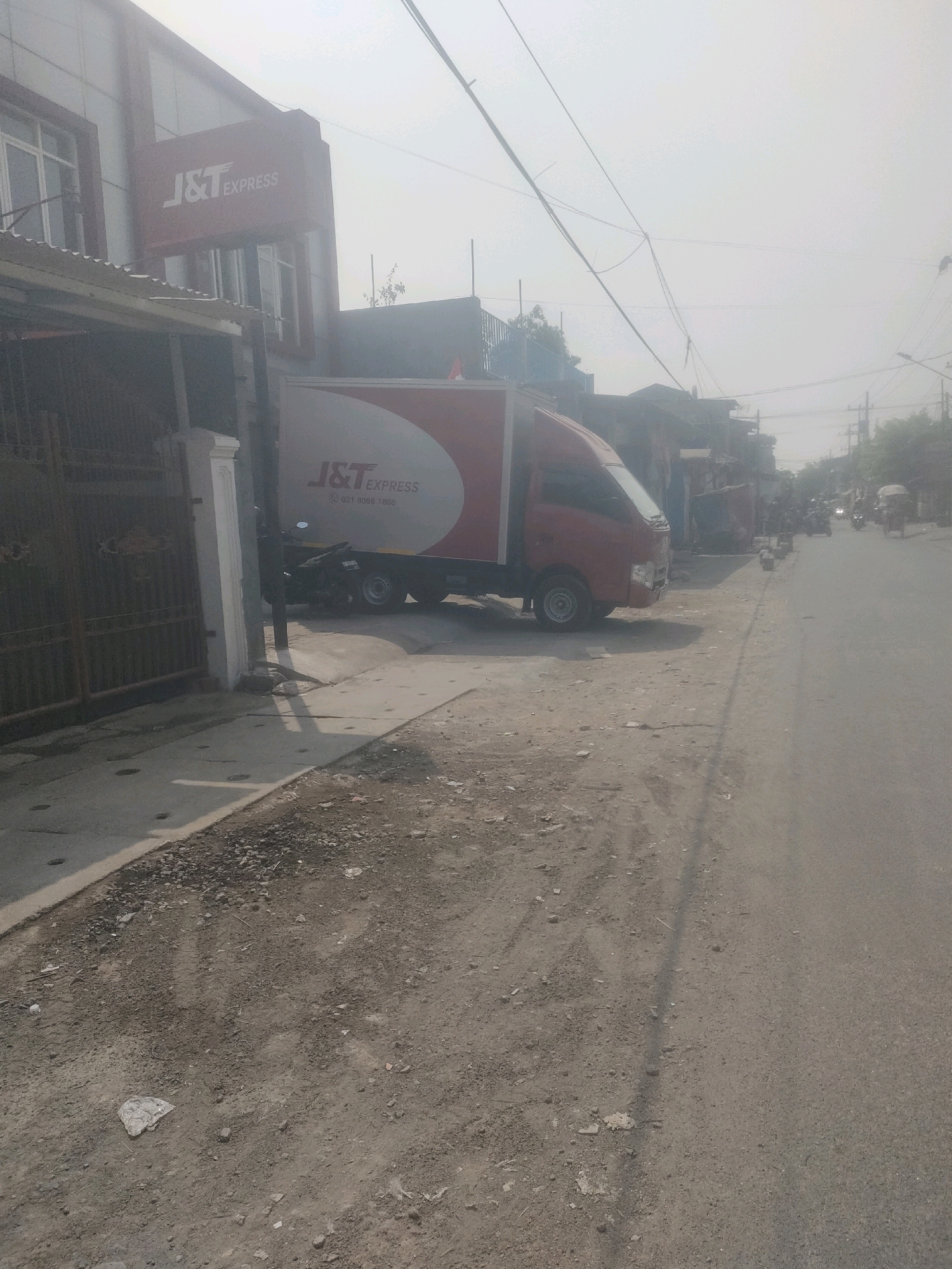 The image depicts a street scene in Surabaya, Indonesia, under a hazy or overcast sky. On the left side of the frame, a red and white J&T Express delivery truck is parked parallel to a building with a J&T Express sign. A motorcycle is visible parked next to the truck, and a metal gate is to the left of the truck. The building appears to be a commercial establishment with windows. The road in front of the truck is unpaved and dusty, with some debris scattered on it. The right side of the image shows a paved road with several motorcycles and a tricycle in the distance, suggesting ongoing traffic. Trees and other buildings line the side of the road. The overall environment appears to be a busy urban or semi-urban area.