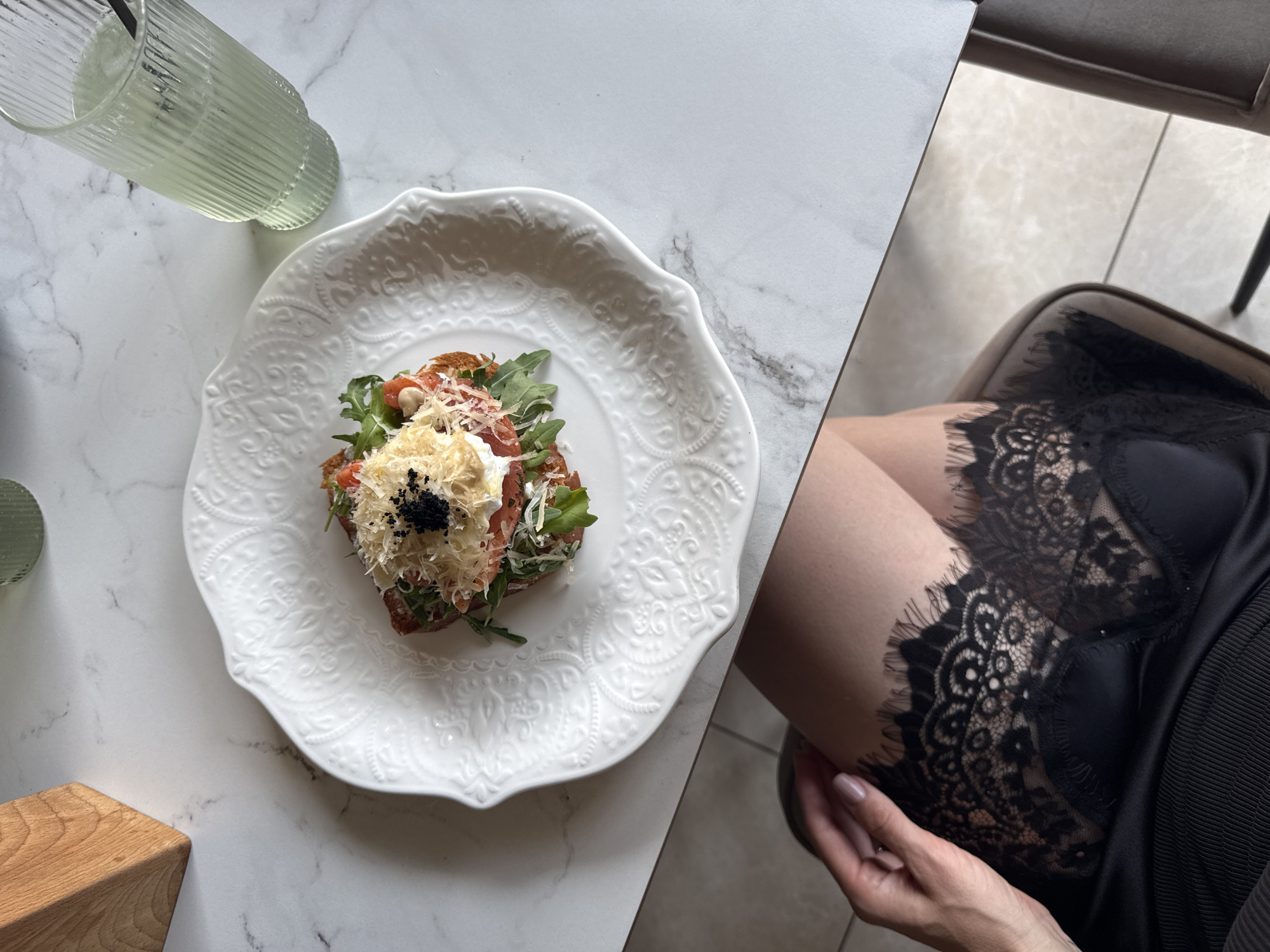 An overhead shot shows a woman's lap, clad in a black lace-trimmed garment, resting on a chair next to a white marble-topped table. On the table sits a white decorative plate with an artistic presentation of food. The dish features a base topped with what appears to be a poached egg, layered with salmon or tomato, and garnished with arugula, shredded cheese, and black caviar. Adjacent to the plate is a tall, fluted glass filled with a pale green beverage and a straw. To the left of the plate, a section of a wooden object, possibly a utensil holder, is visible. The overall setting suggests a dining establishment. No specific geographic indicators pinpoint Ust'-Muny, Russia.
