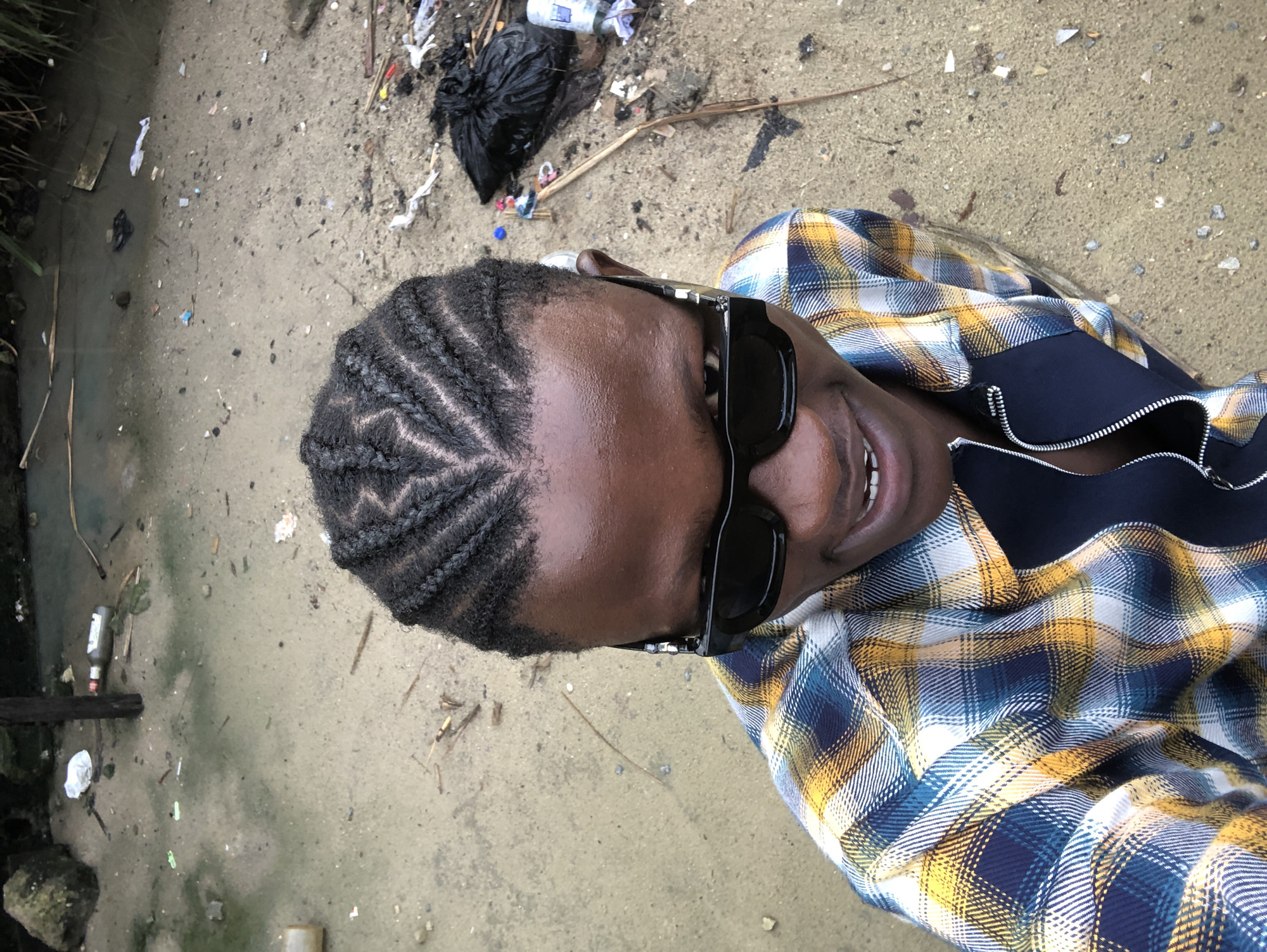 In Ado, Nigeria, a young man with braided hair and black sunglasses poses for a selfie. He is wearing a blue, yellow, and white plaid shirt with a black zipper. The background is a sandy area littered with various debris, including plastic bottles, wrappers, and a black trash bag. The ground appears damp in some areas, suggesting it might be near a body of water.