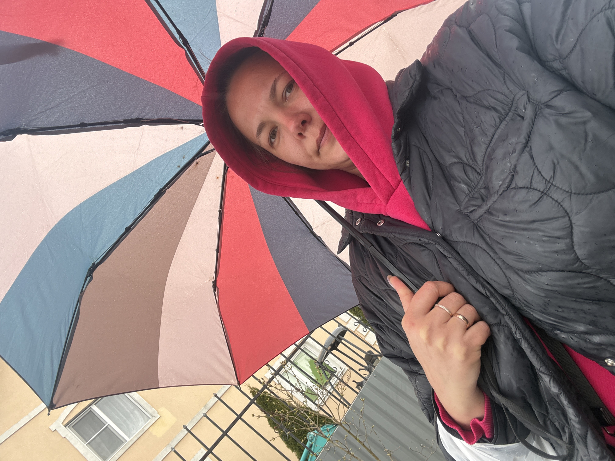 A woman stands under a multi-colored umbrella in what is identified as Dmitrov, Russia. The umbrella features segments of red, white, blue, brown, and grey. The woman wears a red hoodie under a quilted black jacket. Water droplets are visible on the umbrella and jacket, indicating rain. She is looking at the camera. A building with a window is visible in the background, along with a fence, tree, and green container. The woman is holding the edge of her jacket with her right hand, which has two rings on the ring finger.