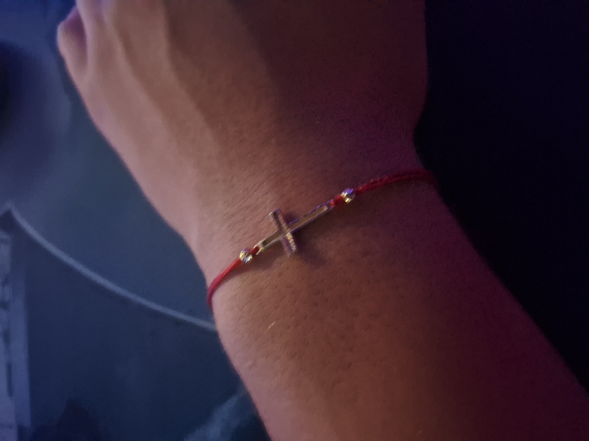 A close-up, angled shot shows a person's left forearm and wrist, wearing a thin red cord bracelet with a small, silver-colored cross pendant. The bracelet is visible against the skin, which has a slightly textured appearance. The background is dark and out of focus, with hints of blue and purple hues, possibly indicating an indoor setting with artificial lighting. The location in Varna, Bulgaria, is not determinable from the image.