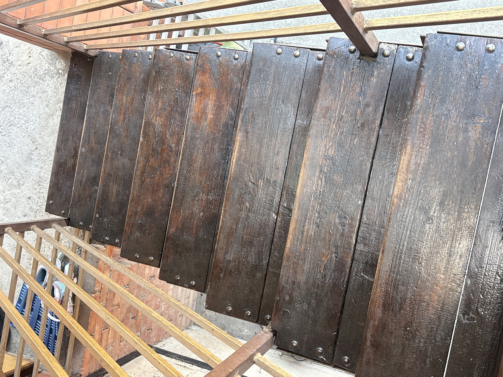 The image shows a set of dark wooden stairs with metal railings, viewed from directly above. There are multiple steps, each made of planks secured with visible metal fasteners. The stairs are positioned outdoors, with a concrete surface visible at the top and a brick wall and metal bars to the left. A pair of athletic shoes and a blue mat are partially visible on the landing at the top of the stairs. The scene depicts an exterior stairway, likely in a residential or public area.