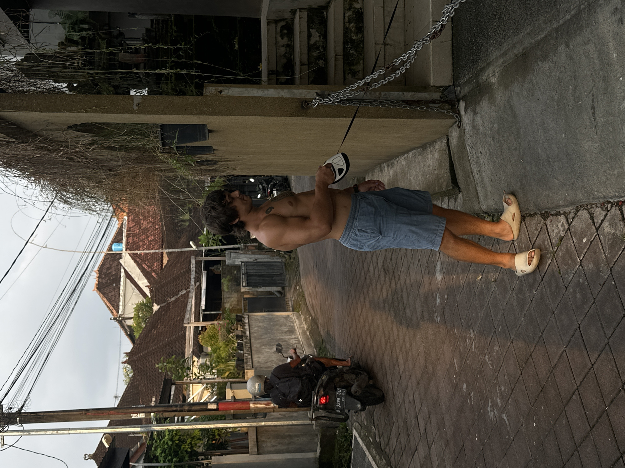 The image depicts a shirtless man with long hair and a tattoo on his chest standing on a paved surface in Dalung, Indonesia. He is wearing blue shorts and sandals, and holding a white and black object with a handle, possibly a measuring tool, that is attached to a chain. The chain is connected to a concrete structure near a set of stairs. In the background, there is a motorcycle parked on the street, along with buildings, power lines, and vegetation typical of a residential area in Indonesia. The image is taken in daylight, suggesting either morning or late afternoon.