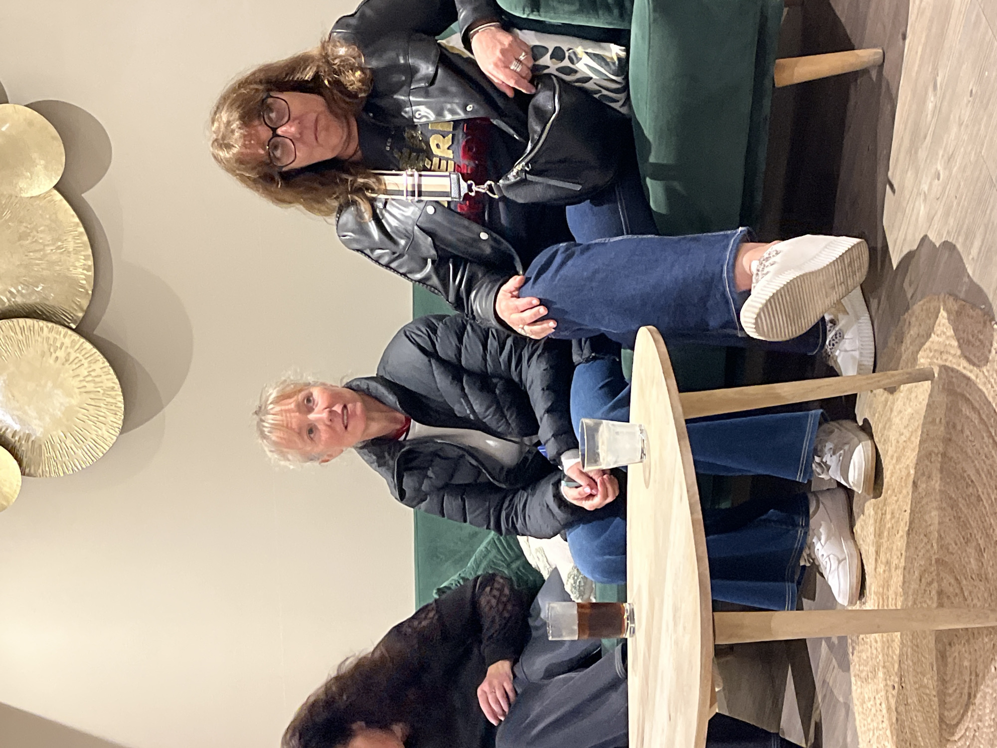 Three women are seated at a round table in what appears to be an indoor lounge area. The woman at the top is wearing glasses, a black leather jacket, and jeans. She has a small black purse over her shoulder. The woman in the middle is wearing a black puffer jacket over a white top, with jeans and white sneakers. The third woman is partially visible, with dark hair and wearing a black dress. They are sitting on a green sofa. Drinks in glasses are on the table. A decorative golden wall hanging is visible on the wall. The table is sitting on a textured mat. This image was taken in Grimaud, France.