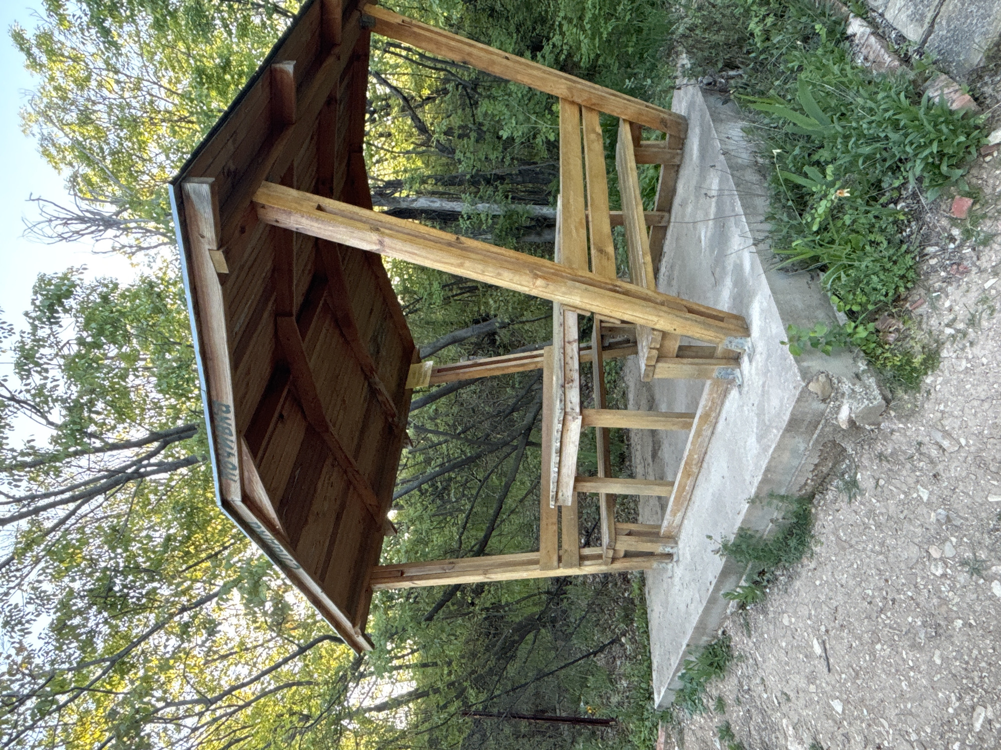 A wooden, open-sided shelter with a pitched roof stands on a concrete foundation in a natural setting. The shelter features wooden beams forming its structure, supporting a roof made of horizontal wooden planks. Inside, there are wooden benches and a table, suggesting a resting or picnic area.
No people or specific activities are depicted; the scene is static. The shelter is nestled among lush green trees with dense foliage, particularly noticeable in the background and through the open sides of the structure. Some bare branches are also visible. The ground around the concrete base is a mix of dirt and gravel, with patches of green weeds and grass.
The lighting indicates daytime, likely late morning or afternoon, with sunlight filtering through the tree canopy. The weather appears clear. A notable detail is faintly visible text on the underside of the roof's overhang, which appears to be Cyrillic. Upon closer inspection, the word "ПОЧИВКА" is discernable, meaning "rest" or "break" in Bulgarian, suggesting the shelter is a designated resting spot, possibly in a park or along a trail in a Bulgarian-speaking region. The overall scene conveys a tranquil, natural environment, offering a place for respite.