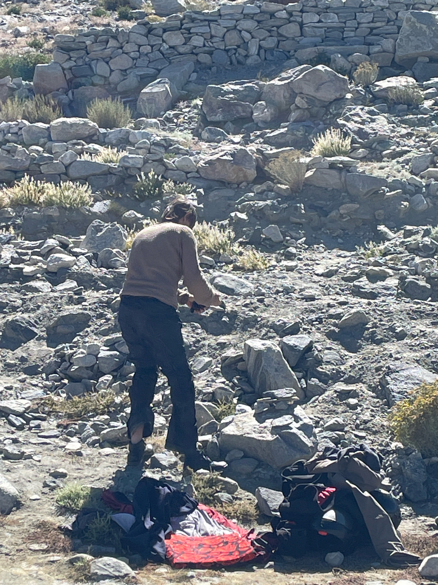 The image depicts a rocky, mountainous terrain with sparse vegetation, characteristic of the high-altitude region near Man, India. In the foreground, a person is standing amidst the rocks, facing away from the camera. They are wearing a light-colored long-sleeved shirt and dark pants. The person appears to be holding a dark object in their right hand. In front of them, a pile of clothing and gear is scattered on the ground, including a red patterned item and dark bags. The background features a stone wall constructed from irregular rocks, possibly remnants of an ancient structure. The overall scene suggests a rugged outdoor environment.