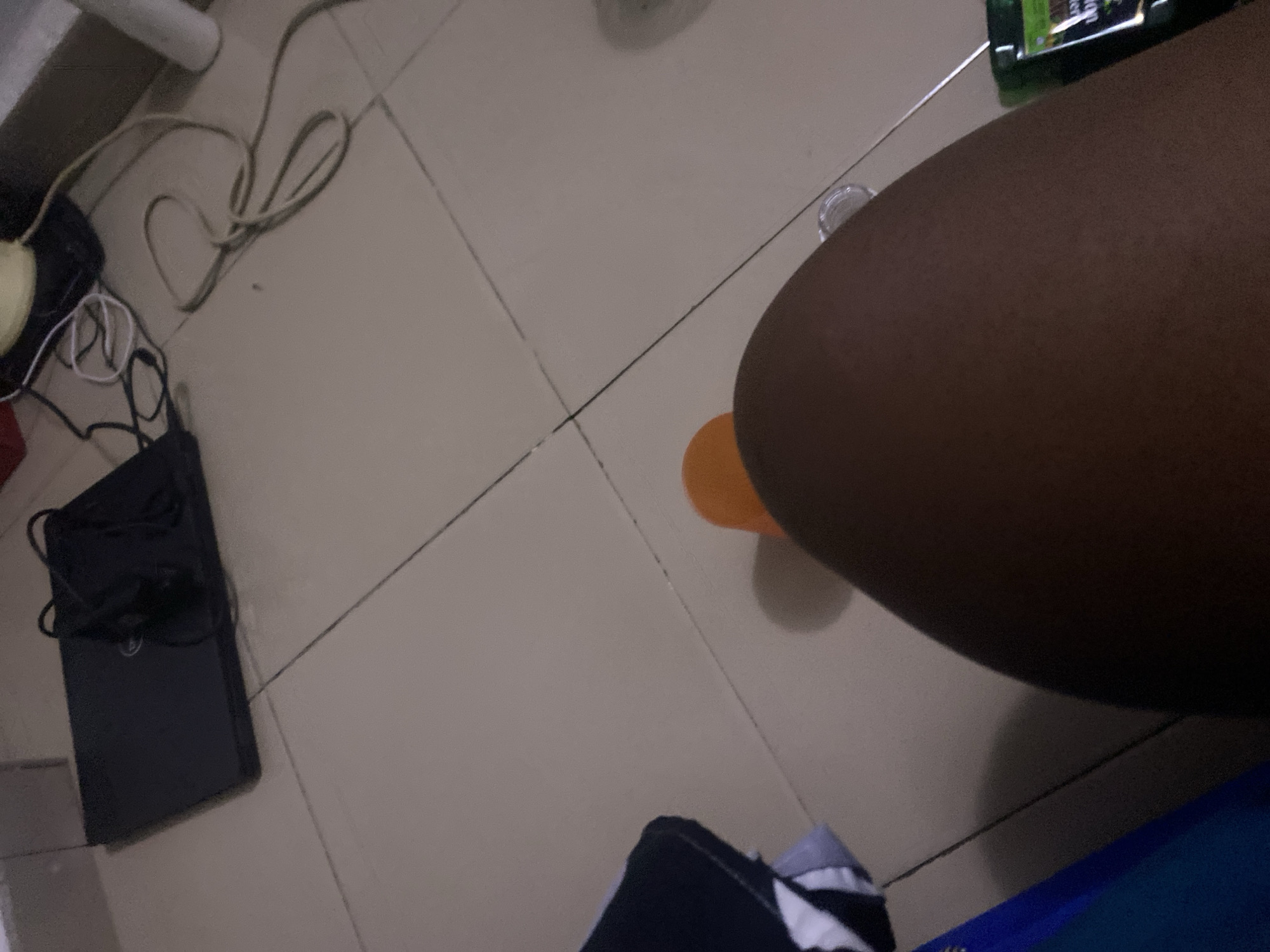 The image shows a tiled floor in what appears to be a residential setting. A dark-colored laptop is visible on the floor in the upper left portion of the image, with its charging cable connected and coiled next to it. Wires and a yellow object, possibly a power adapter, are also present. In the lower half of the image, a person's thigh is in focus, with an orange object, likely a toy or a piece of fruit, resting on it. A dark blue object, possibly a bag or a box, is partially visible on the left side. A green bottle with labels, potentially a cosmetic or cleaning product, is seen on the right side. The overall environment appears to be indoors, with minimal natural light. There is no information in the image to determine a specific location within Howayi, Nigeria.