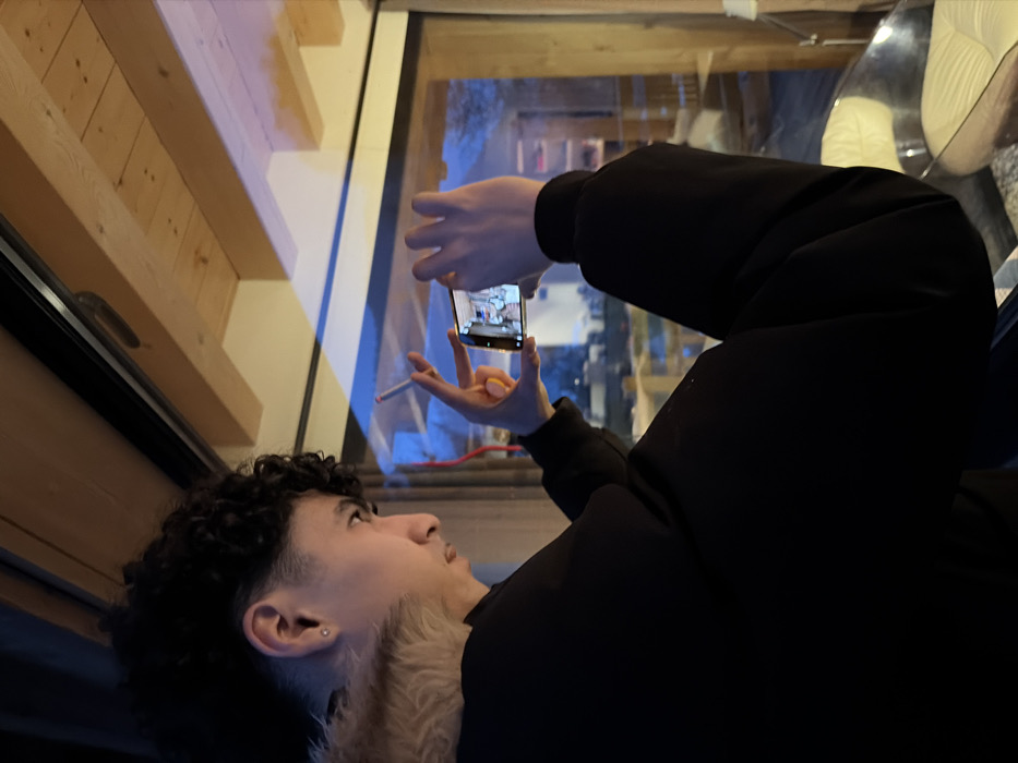 A young individual with dark, curly hair and a faded undercut is depicted in profile, facing right, within an indoor setting in La Clusaz, France. The person wears a black long-sleeved garment with a light-colored, possibly faux fur, collar and a silver stud earring in the visible left earlobe. Both hands grasp a horizontal smartphone, actively capturing or recording an image; its screen displays an interior scene. A lit cigarette is held between the index and middle fingers of the left hand. The background features a large glass window, through which the twilight or night-time landscape of the mountain village of La Clusaz is visible, characterized by distant structures with pitched roofs under artificial illumination. Blue ambient light from the exterior reflects onto interior surfaces. Overhead, exposed wooden ceiling beams are prominent. A white upholstered armchair with a metallic base is partially visible in the lower right foreground.
