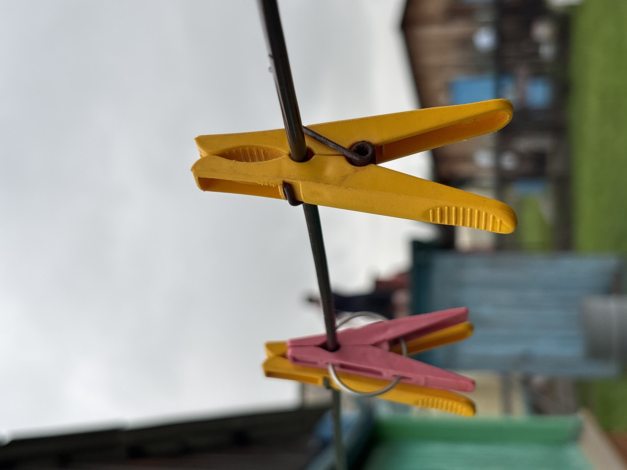 In Gonja, Russia, under a cloudy sky, two clothespins, one pink and one yellow, are clipped to a clothesline. The pink clothespin is on the left, and the yellow one is to its right, slightly overlapping. Behind them, a blurry figure stands near a light blue structure. To the left, a green structure is visible, and in the background, several indistinct wooden buildings stand amidst greenery. The focus is on the clothespins, with the background rendered out of focus.