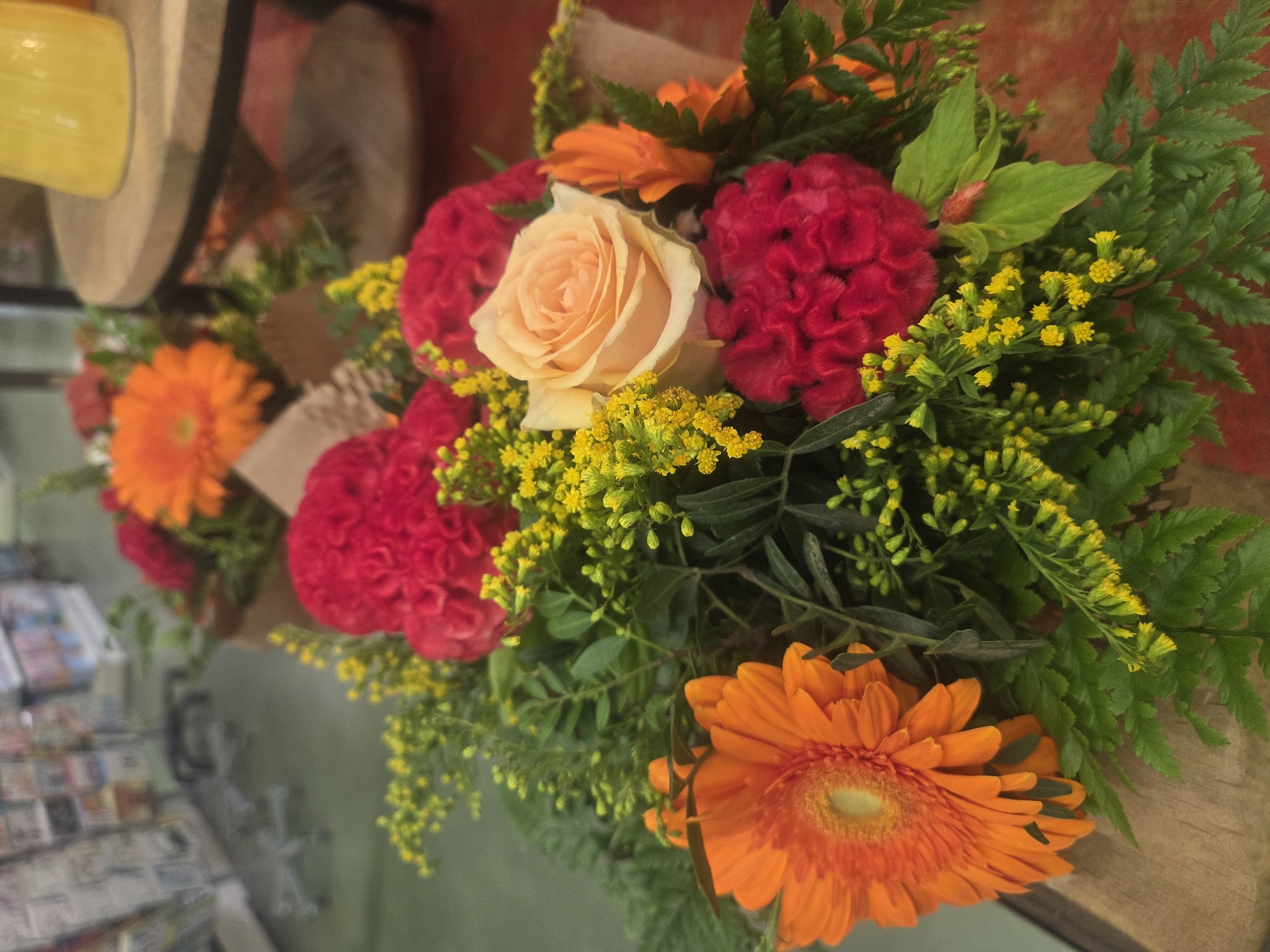 A close-up, eye-level shot captures a vibrant bouquet of flowers, likely a gift or for sale, placed on a wooden surface. The bouquet is a mix of colors and textures, featuring a prominent bright orange gerbera daisy on the left, a soft peachy rose in the center, and several deep red, velvety celosia flowers scattered throughout. Accompanying these larger blooms are delicate sprigs of small yellow flowers and lush green fern-like foliage. The background is blurred, suggesting an indoor setting. To the left, indistinct shelves filled with items hint at a retail environment, possibly a flower shop or market. To the right, a hint of a wooden table and a bright yellow object, potentially a vase or pitcher, are visible. The overall impression is one of freshness and natural beauty, with the bouquet being the clear focal point. The scene is devoid of any people or overt actions, focusing solely on the arrangement of flowers.