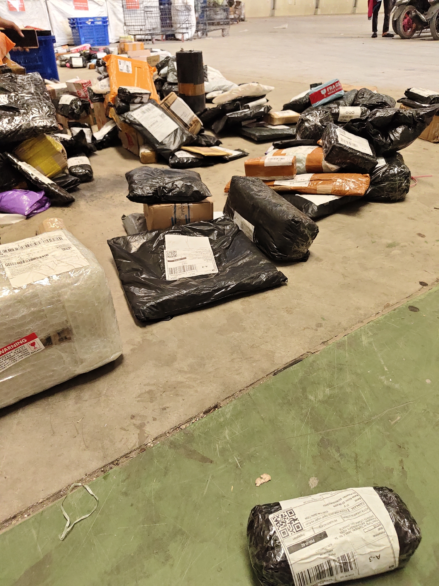 The image depicts a disorganized collection of packages scattered across a concrete floor. The packages vary in size, shape, and packaging material, including bubble wrap, plastic bags, and cardboard boxes. Some packages are wrapped in black plastic, others in brown paper, and a few are encased in clear bubble wrap. Several packages have shipping labels with text and barcodes. In the background, there are stacks of crates, trolleys, and a portion of what appears to be a loading dock or warehouse area. A person is partially visible on the right side of the image, standing near a motorcycle. Another person's hand is visible on the left, holding a mobile phone. The floor is concrete with some areas stained green. The scene suggests a shipping or logistics hub where packages are being processed or have been left in disarray. The mention of "North Jakarta, Indonesia" provides a specific geographical context, implying this scene is likely a part of a distribution center or warehouse within that area.