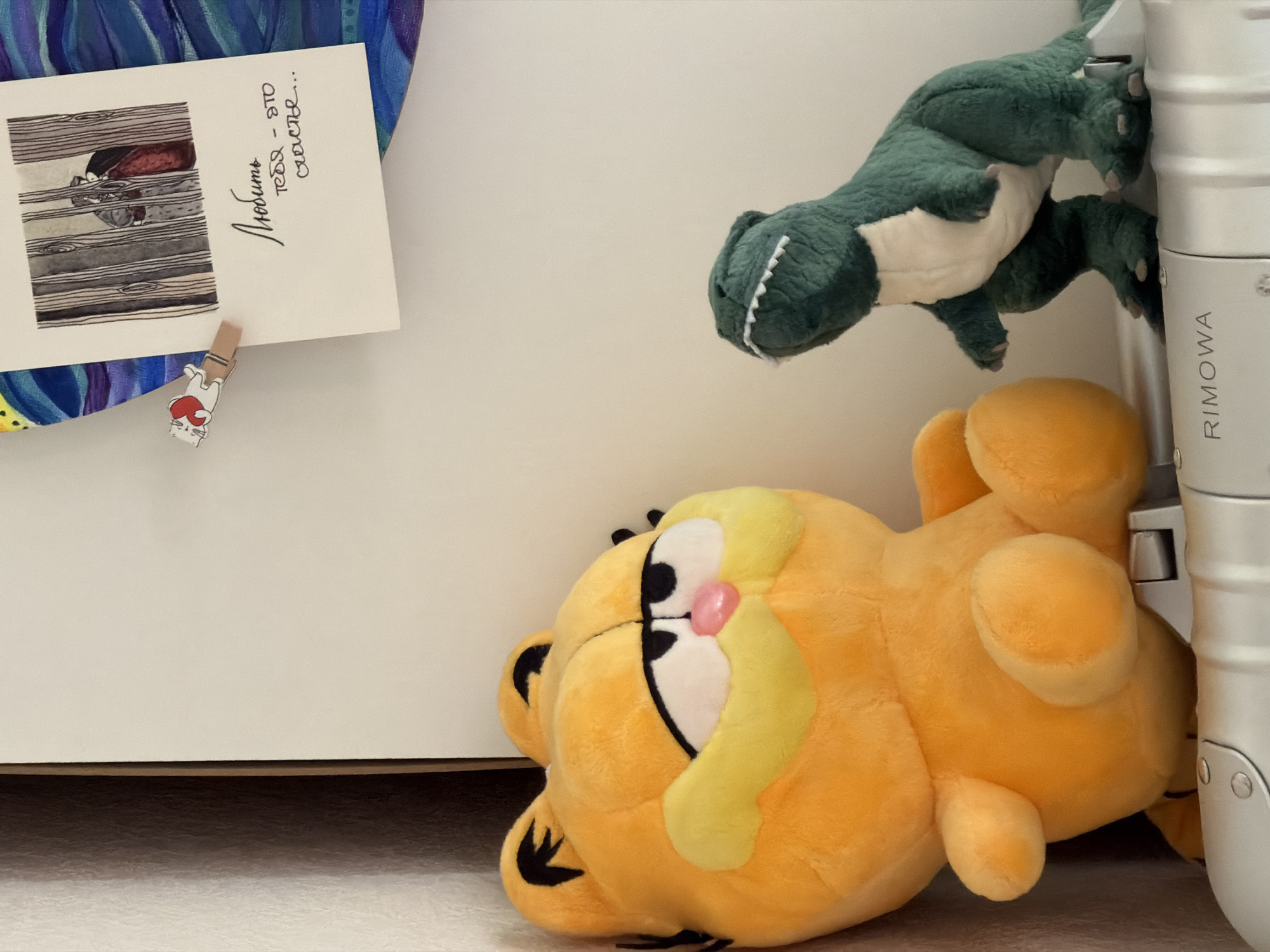 An indoor scene features a playful arrangement of objects against a light-colored wall and floor. In the foreground, an orange Garfield plush toy lies on its back on the light-colored floor, with its head turned to the left, showcasing its distinct eyes and pink nose. Above and to the right, a green dinosaur plush toy, with a white belly and white teeth, clings vertically to the side of a silver metallic object. This object is identifiable as a Rimowa suitcase or similar case, with the brand name "RIMOWA" visibly engraved vertically on its side.
To the left, a white card is fastened to the wall using a small, decorative wooden clothespin shaped like a white cat holding a red heart. The card features an illustration on its left side, depicting what appears to be hands behind a pattern resembling wooden slats or window blinds. On the right side of the card, handwritten text in Russian reads: "Любить тебя - это счастье...", which translates to "To love you is happiness...". Behind the card and extending further left, parts of a vibrant, abstract artwork or fabric in shades of blue and purple are visible. The scene is well-lit, suggesting daytime or bright indoor lighting, creating a warm and personal atmosphere. The Russian text on the card indicates a Russian-speaking context.