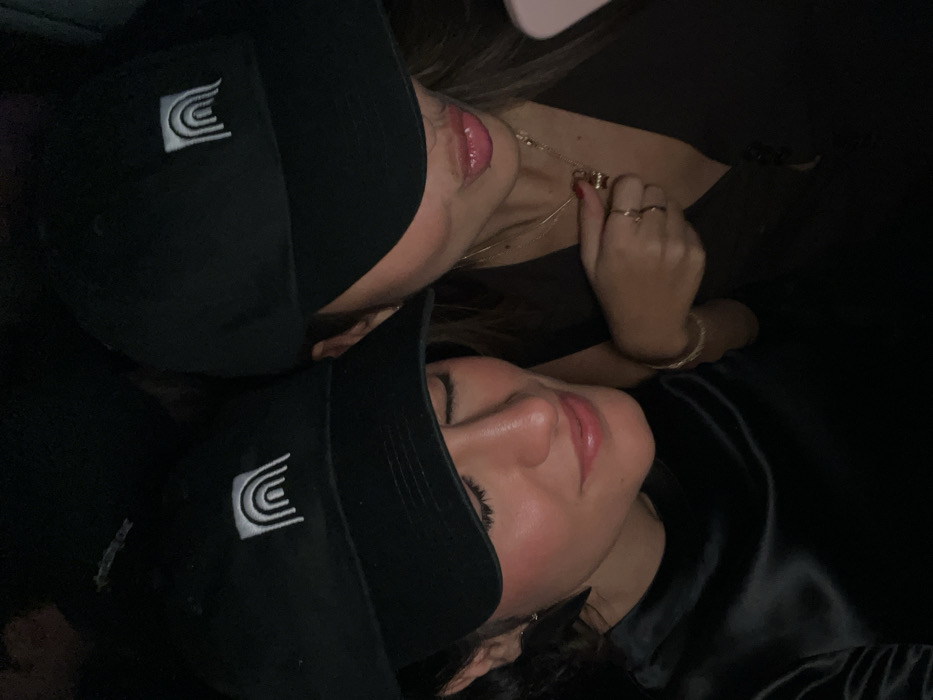 Two women wearing black baseball caps with a white logo are close together. The lighting is dark, and the focus is on their faces. One woman wears a necklace and rings. The other woman's attire appears to be a shiny black fabric. Both have glossy lips.
