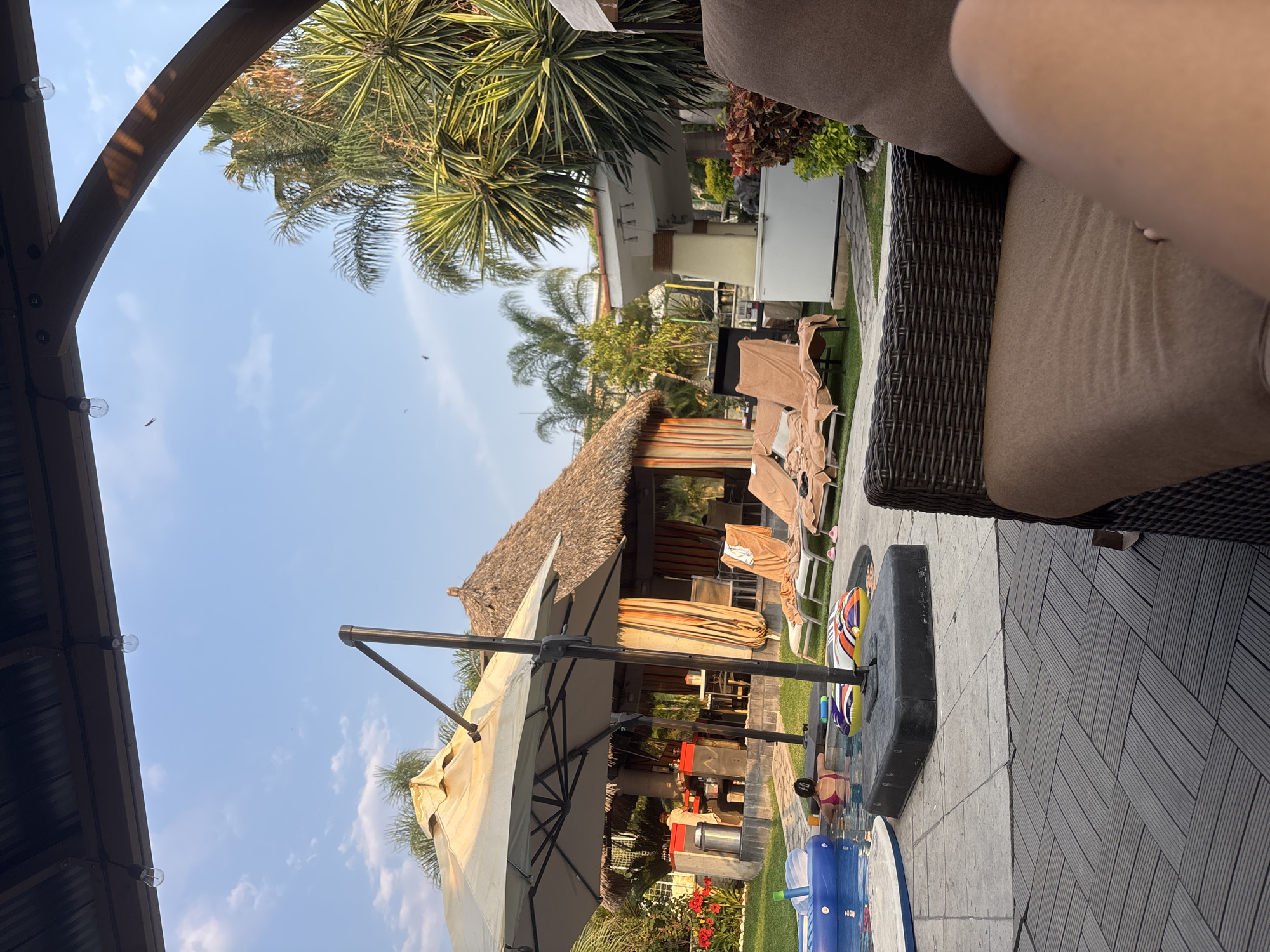 The image shows an outdoor scene, possibly in a backyard, on a sunny day. The perspective is from a seated position on a patio chair with woven rattan framing.
Visible elements include:
* A structure with a thatched roof.
* Lounge chairs with towels draped over them.
* A large patio umbrella.
* Various plants and trees, including palm trees.
* Pool toys.
* A partial view of a building in the background.
* The flooring consists of wooden decking.
* Some structures are on an elevated part of the yard.