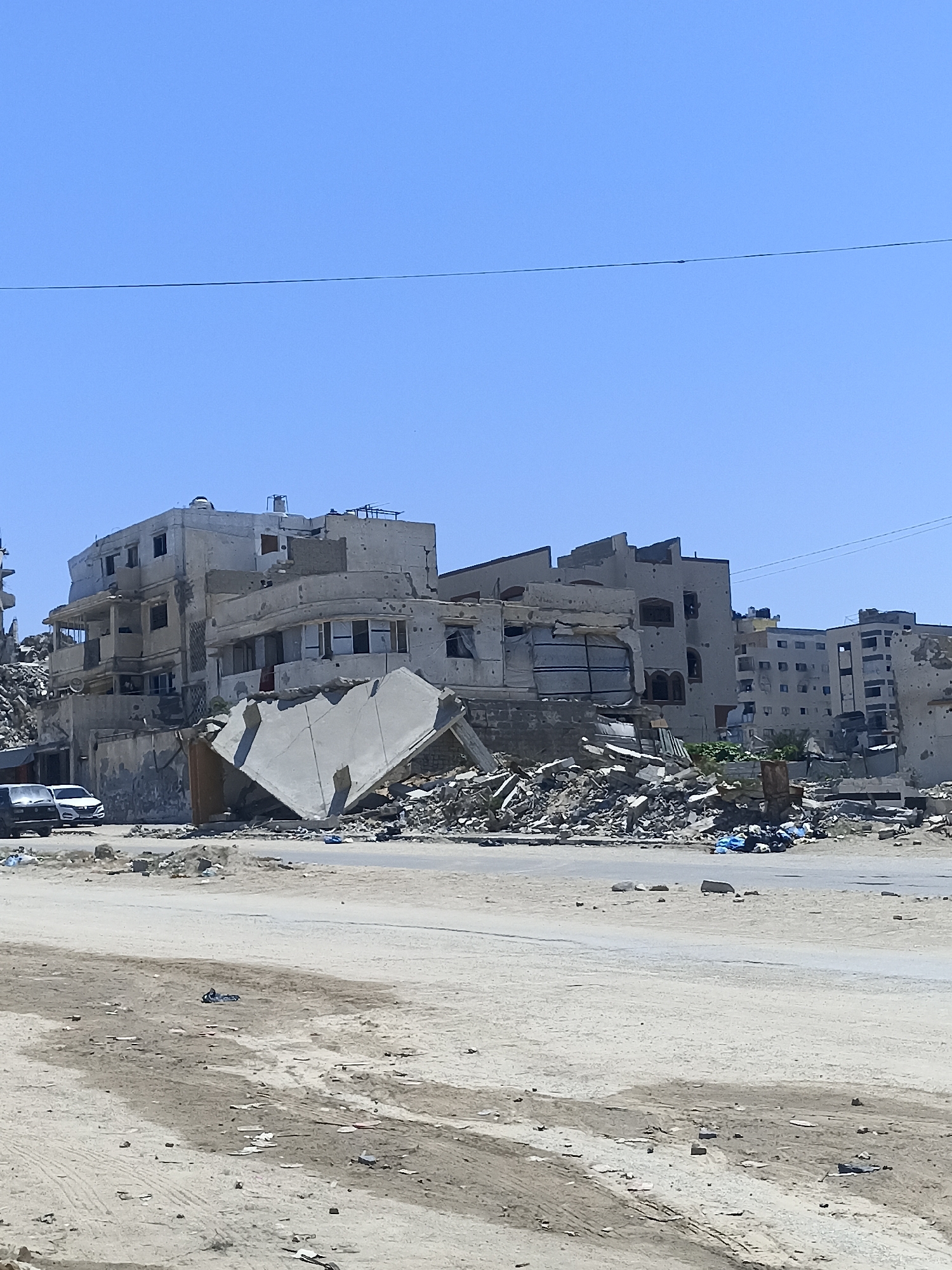 The image depicts a street scene in Gaza, showing heavily damaged buildings under a clear blue sky. The structures are in a state of ruin, with debris scattered across the ground. Several cars are parked on the left side of the road. The foreground is a sandy, unpaved area with tire tracks and scattered debris. The overall impression is one of devastation.