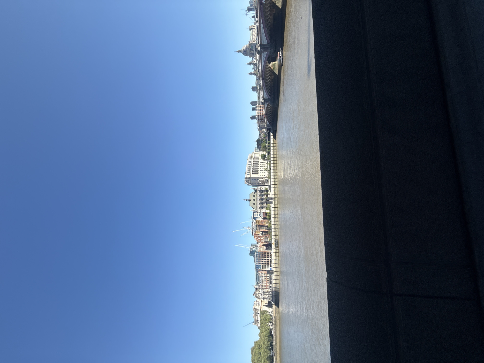 This is an eye-level, outdoor shot, taken from a window or high vantage point, of the London skyline. The image is oriented vertically. On the left is a clear, blue sky. A muddy river runs horizontally through the center of the image with buildings on the far bank. In the upper center, Saint Paul's Cathedral can be seen. There are a number of white and grey buildings lining the river with construction cranes dotted along the skyline. The right side of the image shows a dark, solid structure, possibly a wall.