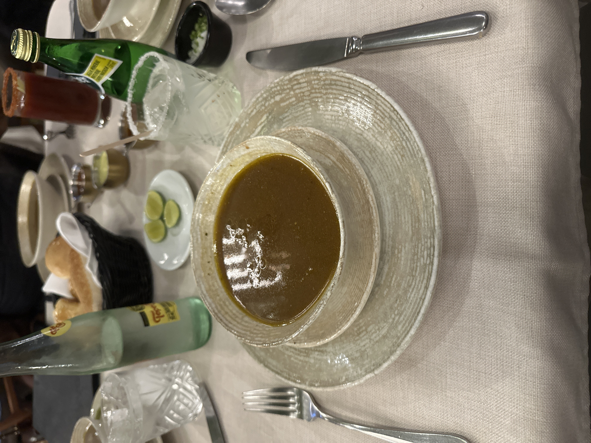 The image depicts a table setting, likely at a restaurant, featuring a bowl of soup placed on a saucer and a larger plate. Utensils, including a knife and fork, are positioned beside the plates. Other items on the table include drinks, such as Topo Chico mineral water and what appears to be a Mexican cocktail (possibly a Michelada), as well as a small bowl containing sliced limes. There is also a bread basket in the periphery. Based on the overall scene, it is plausible that the image was taken in San Bernardino Tlaxcalancingo, Mexico, considering the typical dishes and drinks found within the region.