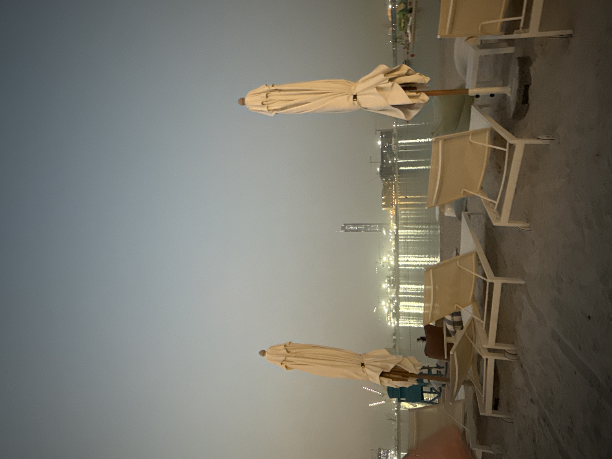 The image depicts a beach scene at dusk in an unspecified location within the United Arab Emirates. In the foreground, several empty lounge chairs are arranged on the sand. Two closed beach umbrellas are visible, one on the left and another on the right side of the frame. Beyond the beach, a body of water reflects the lights of a city skyline. Several buildings, including a tall skyscraper and construction cranes, are silhouetted against the twilight sky. The overall atmosphere is tranquil, with the subdued lighting suggesting the end of the day.