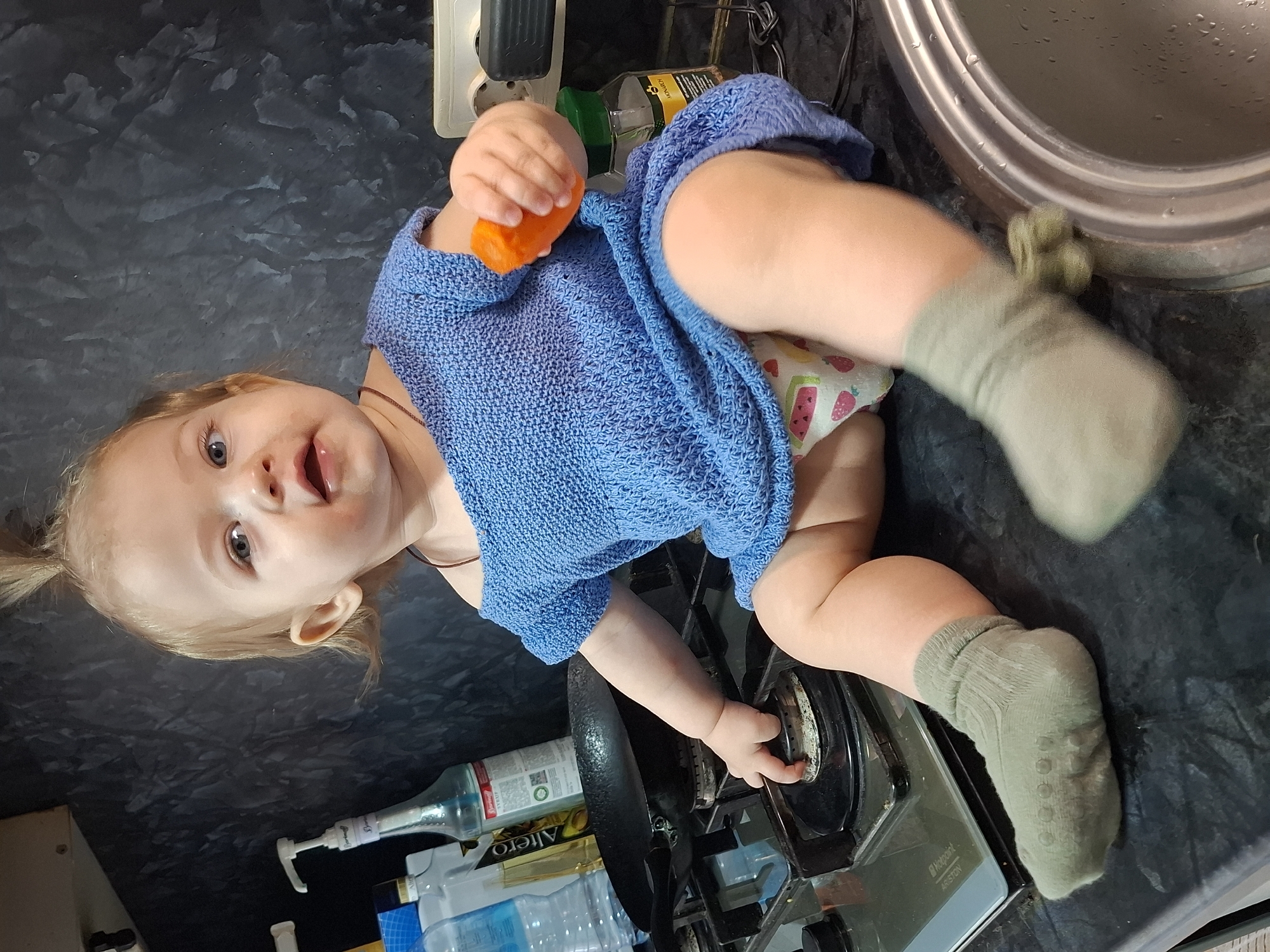 A baby in a blue knitted sweater and socks, is lying on a black countertop, likely in a kitchen. The baby is holding an orange toy in one hand and reaching for a gas burner on a stove with the other. Bottles and cleaning supplies are visible near the stove. A metal sink is nearby. The image was taken indoors. The location of the image is Chaikovsky, Russia.
