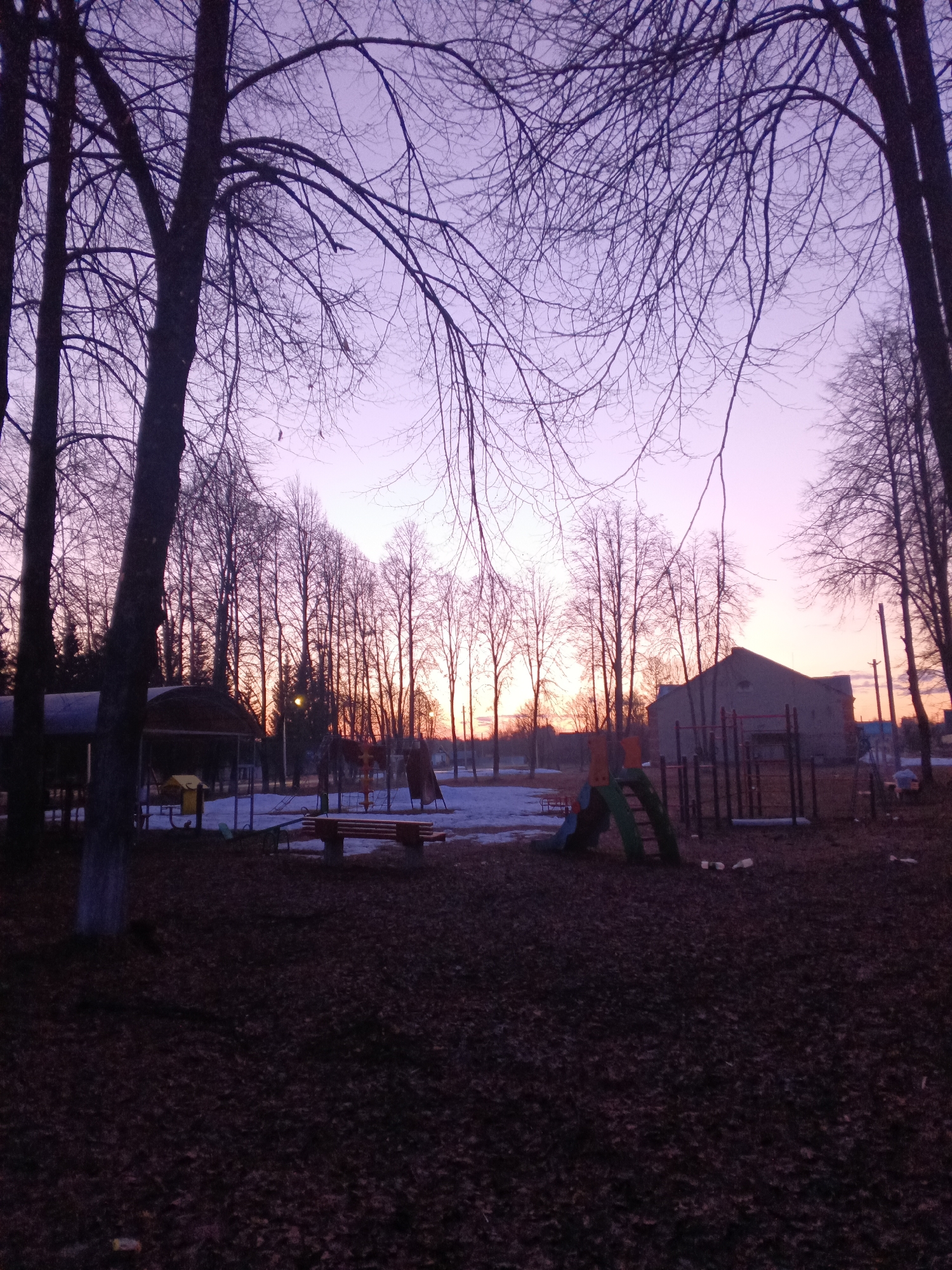 The image depicts a playground in Krasny Mayak, Russia, during dusk or dawn. The sky is a mix of purples and oranges. Bare trees frame the scene. A playground with a slide, swings, and a bench are visible. Remnants of snow are on the ground. The ground is covered in fallen leaves. In the background is a building. No people are visible.