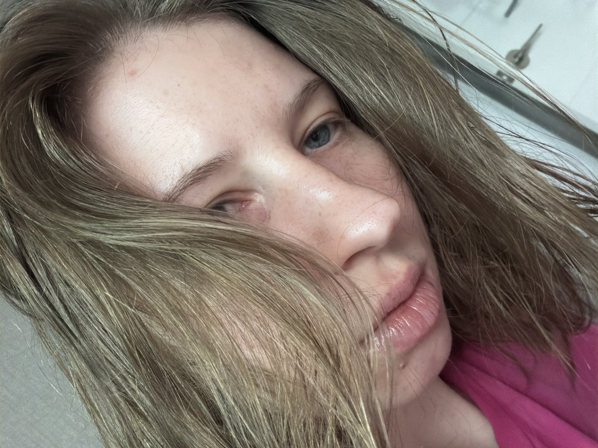 A close-up, angled shot shows the face of a young woman with light brown hair. Her hair is draped across her face, obscuring part of her forehead and eye. She is wearing a bright pink top. The background appears to be a neutral, light-colored wall, with a hint of what might be a door or cabinet on the right side. The lighting is soft and natural. No specific location in Dongducheon-si, South Korea can be determined from the image.