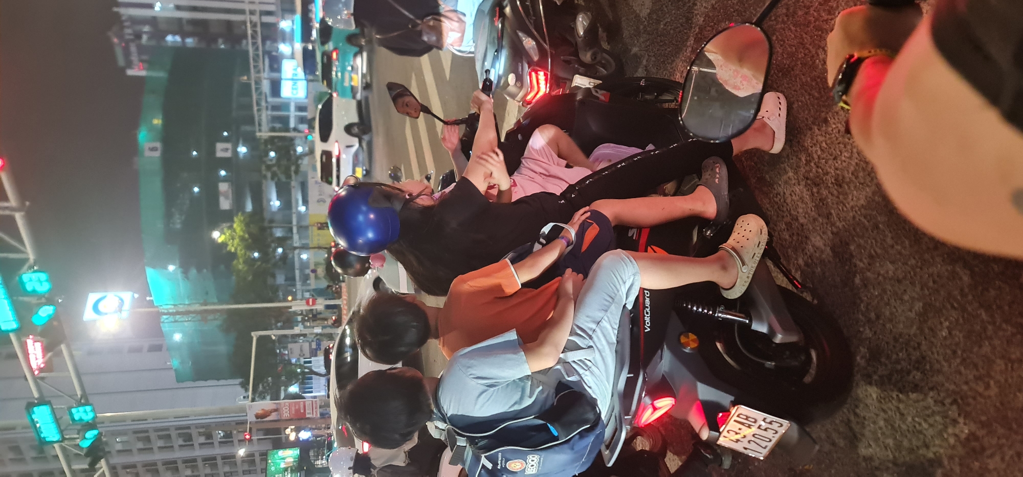 Scene: A traffic stop in Đà Nẵng, Vietnam at night.
People: Four individuals of Asian descent are grouped around a scooter. One wears a blue helmet. The other three are not wearing helmets.
Subjects: A scooter. Cars are visible in the background.
Objects: The scooter is black with orange and silver decals. It has a license plate that reads "43-AB 120.55". The people are wearing casual clothing. The person in the foreground is wearing an orange shirt and light blue shorts.
Actions: The individuals are gathered around the scooter. The person on the scooter appears to be looking in the mirror.
Interactions: The individuals are positioned in a close group.
Environment: The scene takes place at night in an urban environment. Streetlights and traffic lights illuminate the scene. Buildings are visible in the background.