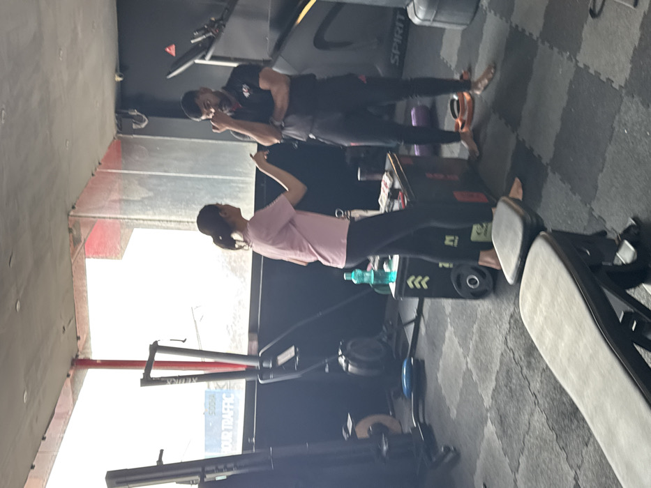 A woman in a pink shirt and black leggings stretches, seemingly in a gym. Behind her is gym equipment including a rowing machine and a squat box. A man in black workout clothes stands near a treadmill, looking at his phone. The gym has a black wall and tiled flooring.