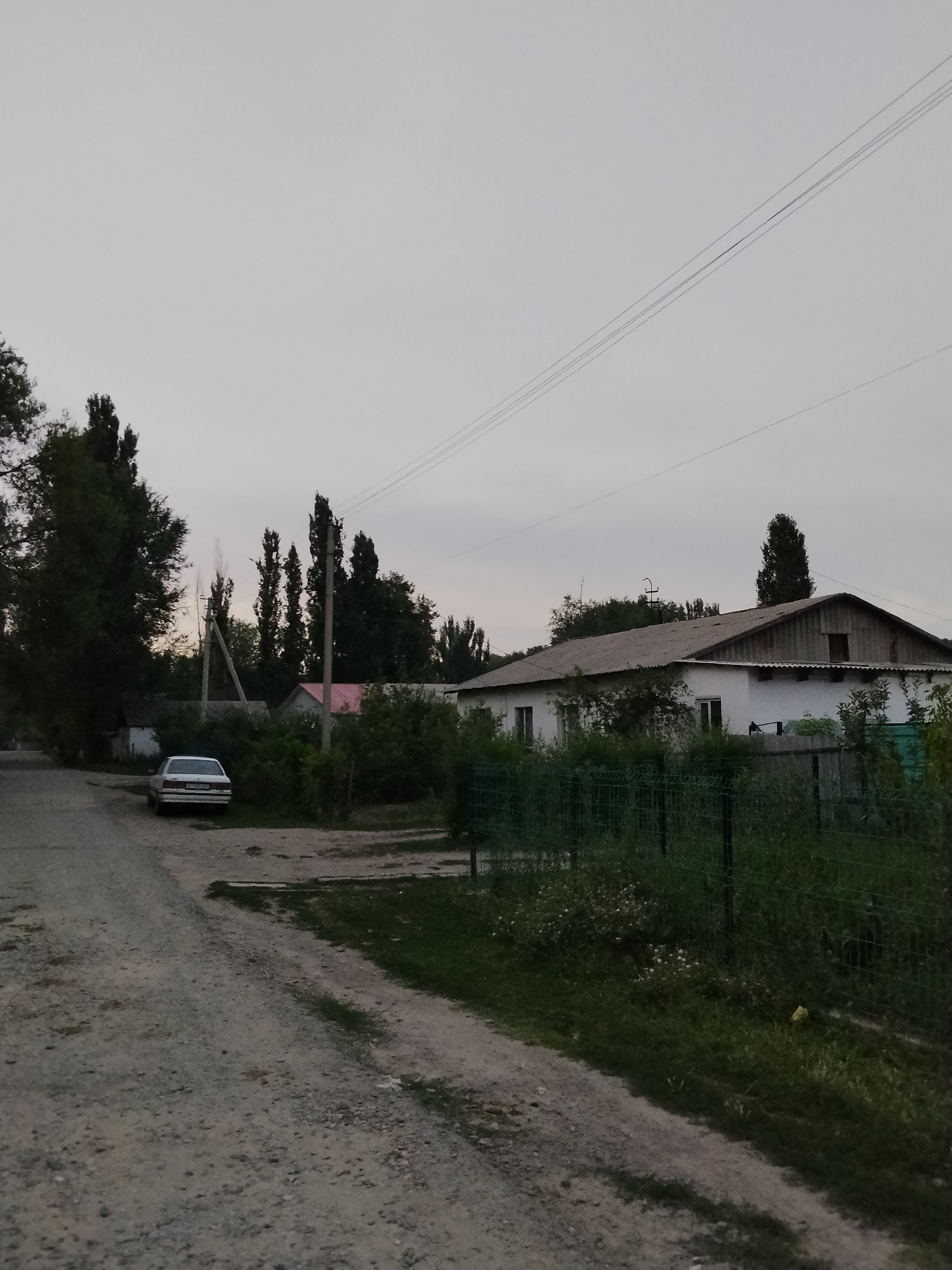 The image depicts a rural street scene in the city of Djany-Pahta, Kyrgyzstan. A dirt road occupies the foreground, leading towards a cluster of buildings and trees in the background. A white sedan is parked on the left side of the road, facing away from the camera. Behind the car, a white building with a gray roof is visible, partially obscured by dense green foliage. Further back, a longer, white, barn-like structure with a weathered gray roof spans across the mid-ground. The sky above is overcast and pale. Power lines are visible diagonally across the upper portion of the image. The environment suggests a quiet, semi-rural setting.