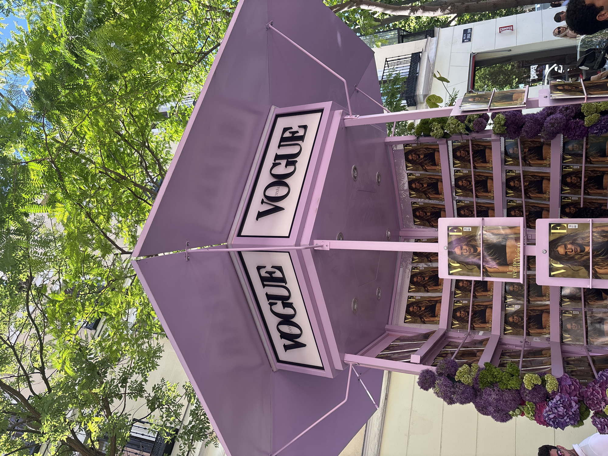The image depicts a street scene in Madrid, Spain, featuring a promotional structure for Vogue magazine. The structure is predominantly lavender in color and has the Vogue logo displayed prominently on two sides. Numerous copies of a Vogue magazine, featuring a woman on the cover, are arranged on shelves around the structure. Purple and green flowers are used as decorative elements. The background includes trees and buildings, with people visible in the distance, some of whom appear to be looking at the Vogue display.