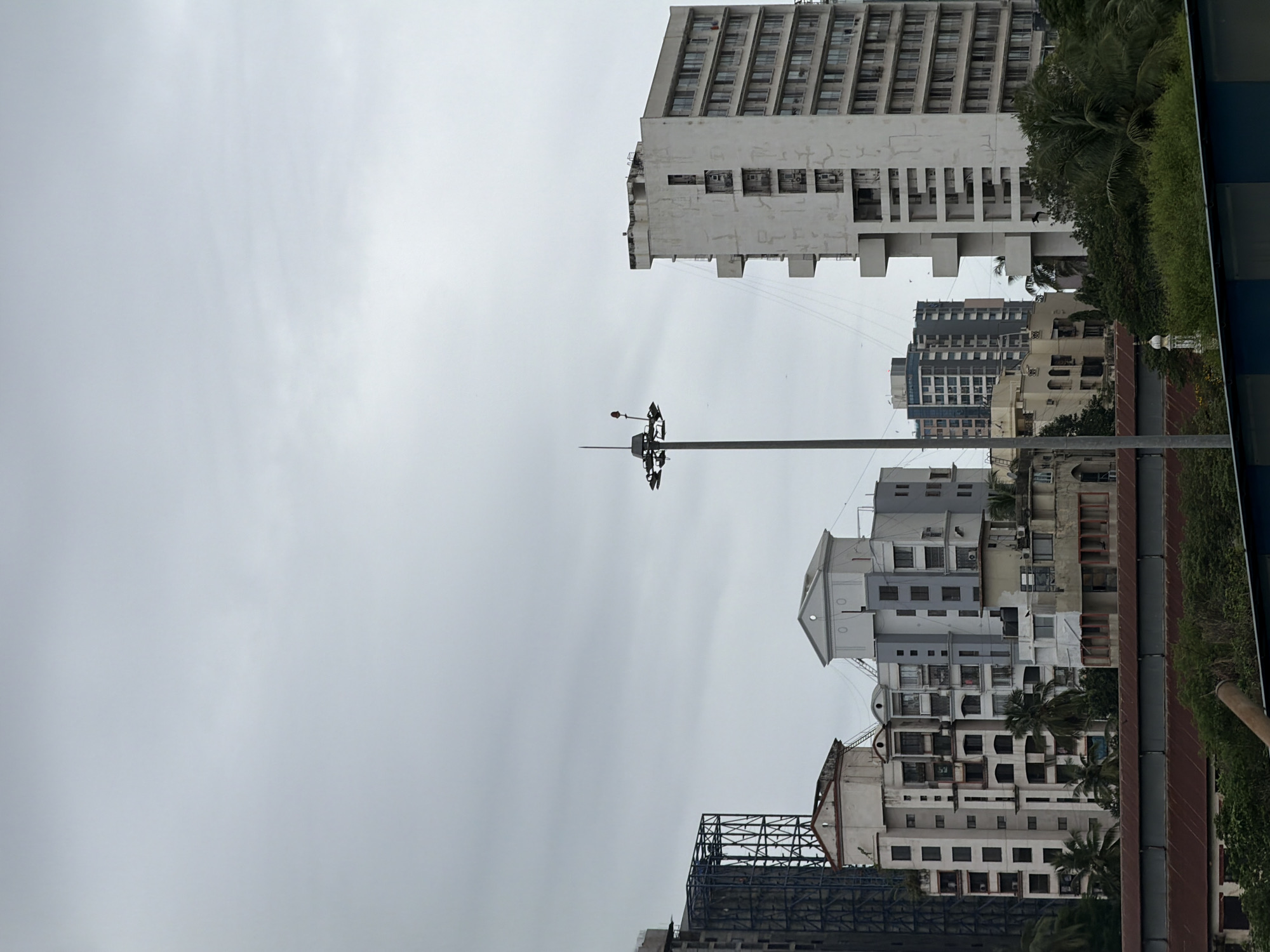 The image depicts a cityscape of Mumbai, India, under an overcast sky. In the foreground, a reddish-brown metal roof structure runs horizontally across the lower portion of the frame, partially obscured by lush green foliage. Behind this, a series of apartment buildings of varying heights and architectural styles dominate the mid-ground.
To the left, a tall, dark, unfinished building with visible scaffolding stands beside a lighter-colored building with a distinctive arched facade. Adjacent to these are a cluster of cream and light gray multi-story residential buildings, featuring numerous windows and balconies. Among them, a tall, white, modern apartment building with a grid-like facade and cantilevered sections is prominent on the right side of the frame. Several palm trees and other tropical vegetation are visible peeking out from between the buildings.
A tall, dark lamppost with a lighting fixture is positioned in the center of the image, extending upwards into the sky. The sky itself is a uniform pale gray, suggesting cloudy or potentially rainy weather. In the far distance, a few more modern high-rise buildings are visible. A minaret of a mosque is subtly visible amidst the trees and buildings on the right side of the frame, a common sight in Mumbai. The overall impression is one of urban density with a mix of older and newer construction.