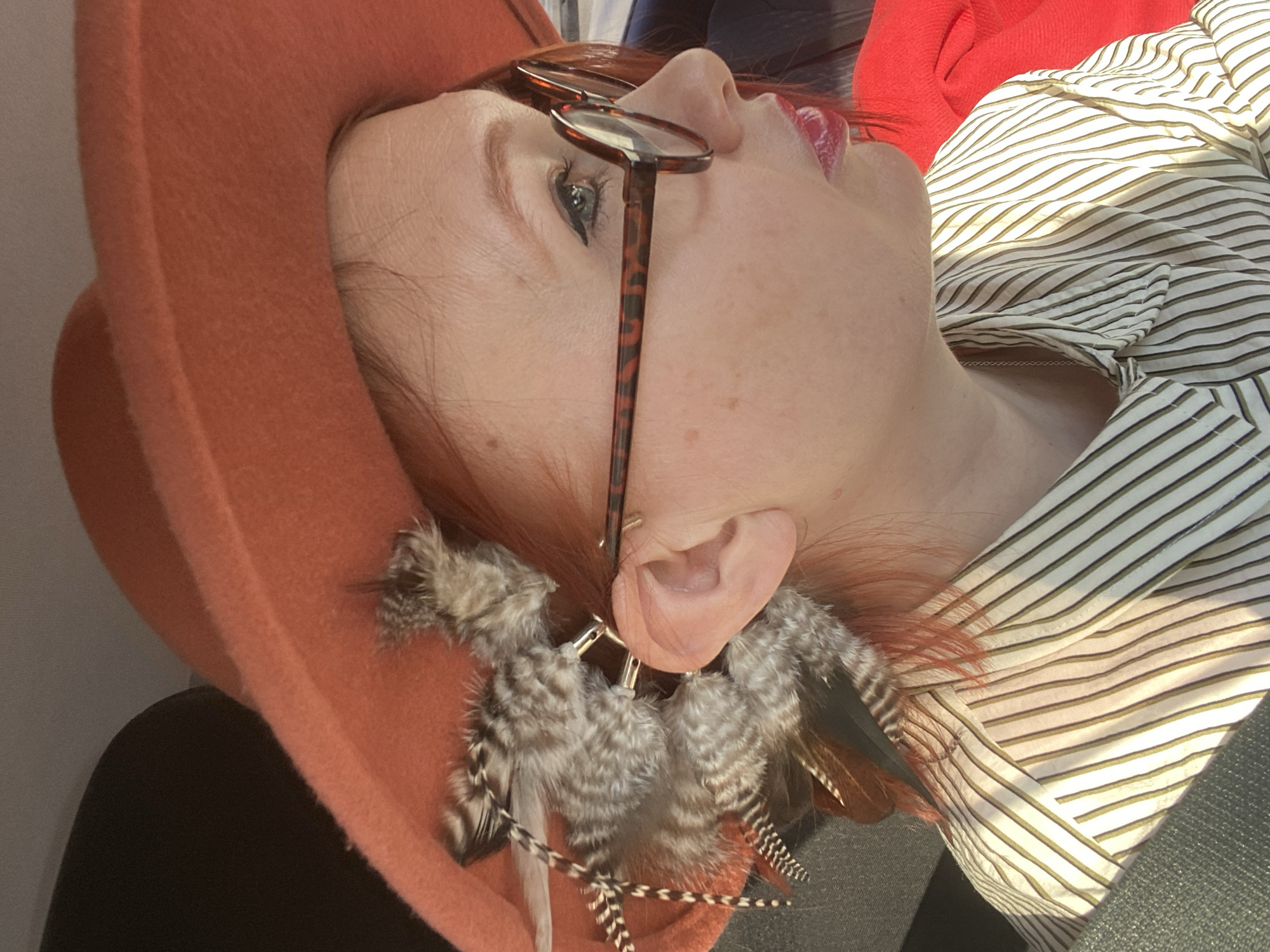 This close-up portrait, taken at an upward angle, features a person, likely female, from the neck up, reclined and looking towards the top right of the frame. The individual has fair skin, reddish-brown hair, and is wearing dark eyeliner and bright red lipstick.
Prominently featured are a pair of tortoise-shell framed eyeglasses, resting on the nose with lenses partially covering one eye. The person is adorned with large, multi-feathered earrings in their earlobes, featuring grey, brown, and speckled feathers attached to gold-colored rings. A delicate silver chain is visible around the neck.
The person is wearing a wide-brimmed hat in a reddish-orange or terracotta hue, which frames the left side of their head. A white shirt with thin, dark (possibly olive green) stripes is visible on the torso, with a hint of a red garment behind the head and a dark garment in the lower left.
The setting appears to be indoors, with a light, plain surface behind the head and a darker, textured surface in the bottom right. The lighting suggests natural daylight, casting bright highlights on the striped shirt and the person's skin. There is no specific activity taking place beyond the person relaxing or posing. No visible text is present in the image. The provided location context is Yekaterinburg, Russia.