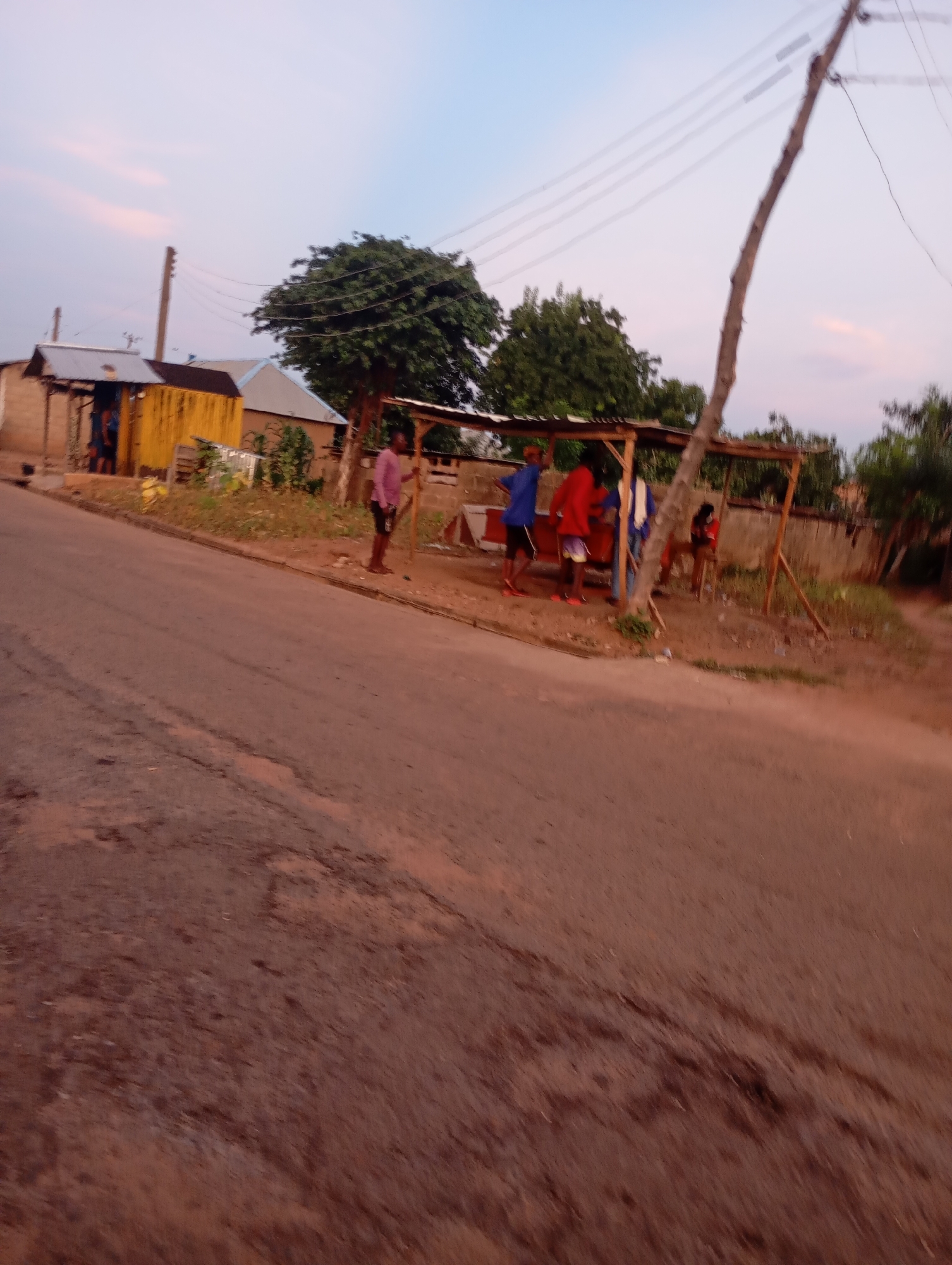 The image shows a roadside scene in Shendam, Nigeria, likely during the early morning or late evening given the soft lighting. A paved road occupies the foreground, with a dirt shoulder on the right. A cluster of informal structures, possibly market stalls or small businesses, are situated along the roadside. These structures have corrugated metal roofs and wooden supports. Several people are present, appearing to be interacting around these stalls. One individual in the foreground, on the right side of the road, is wearing a pink shirt and dark shorts and appears to be holding a stick. Other individuals are clustered under the shaded structure, some wearing t-shirts and shorts. Trees with dense foliage are visible behind the structures, and a utility pole with overhead wires stands to the left of the frame. The overall atmosphere suggests a common, everyday scene in a Nigerian town.